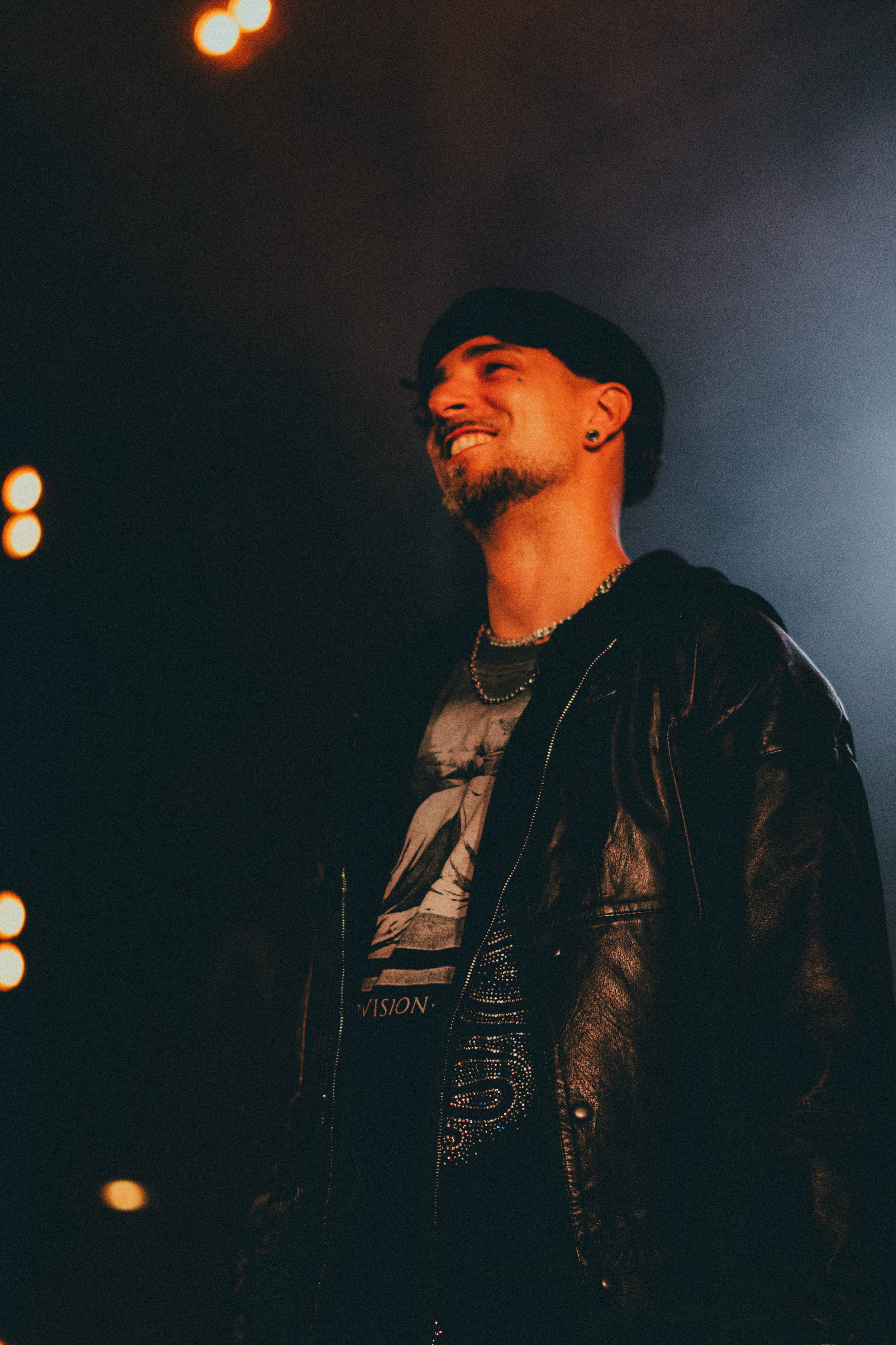 Un homme avec une barbe, portant une casquette noire, des boucles d'oreilles et un collier, souriant dans un environnement sombre avec des lumières chaudes derrière lui.