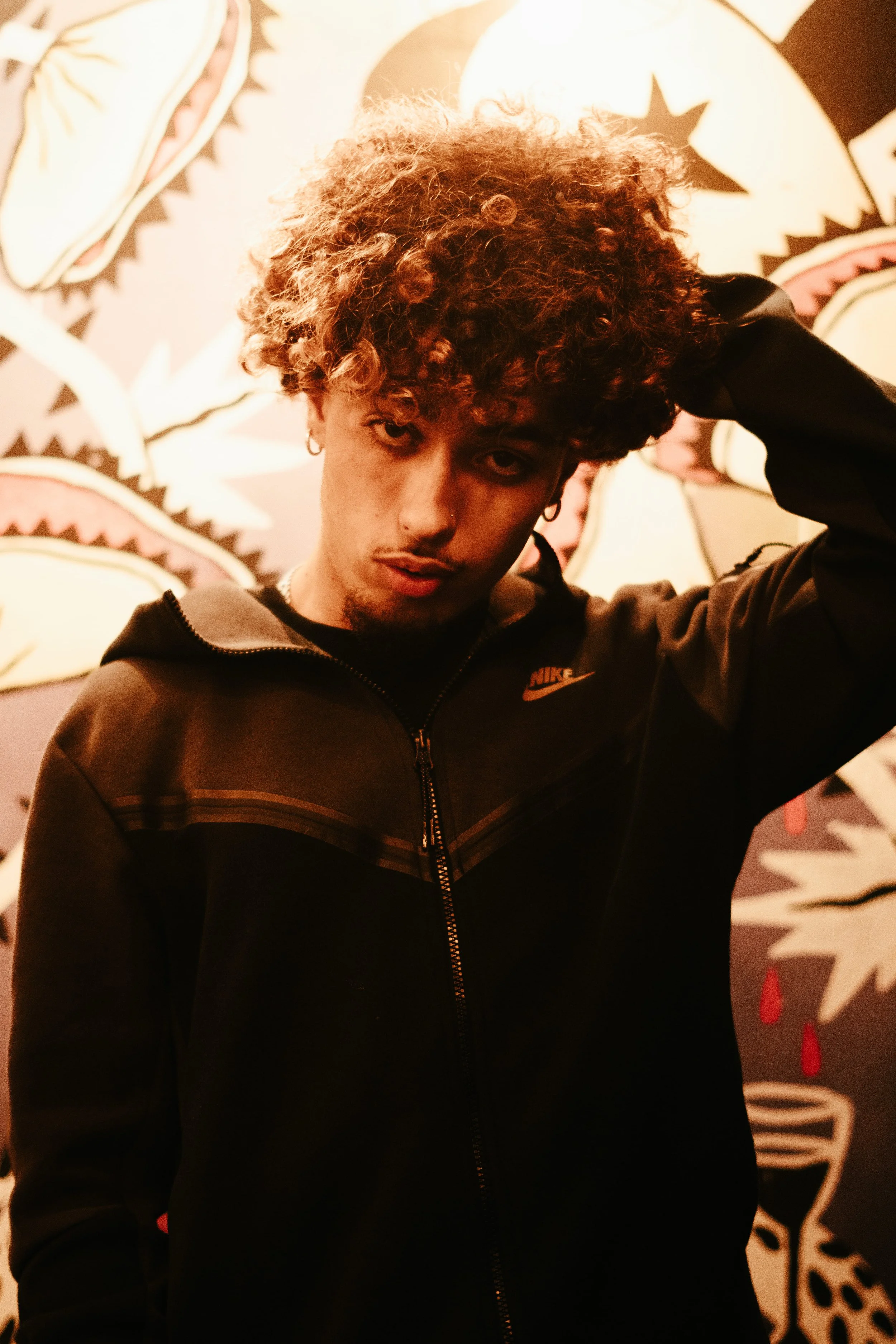 Jeune homme avec cheveux bouclés portant une veste noire Nike, posant devant un mur avec un dessin artistique coloré.