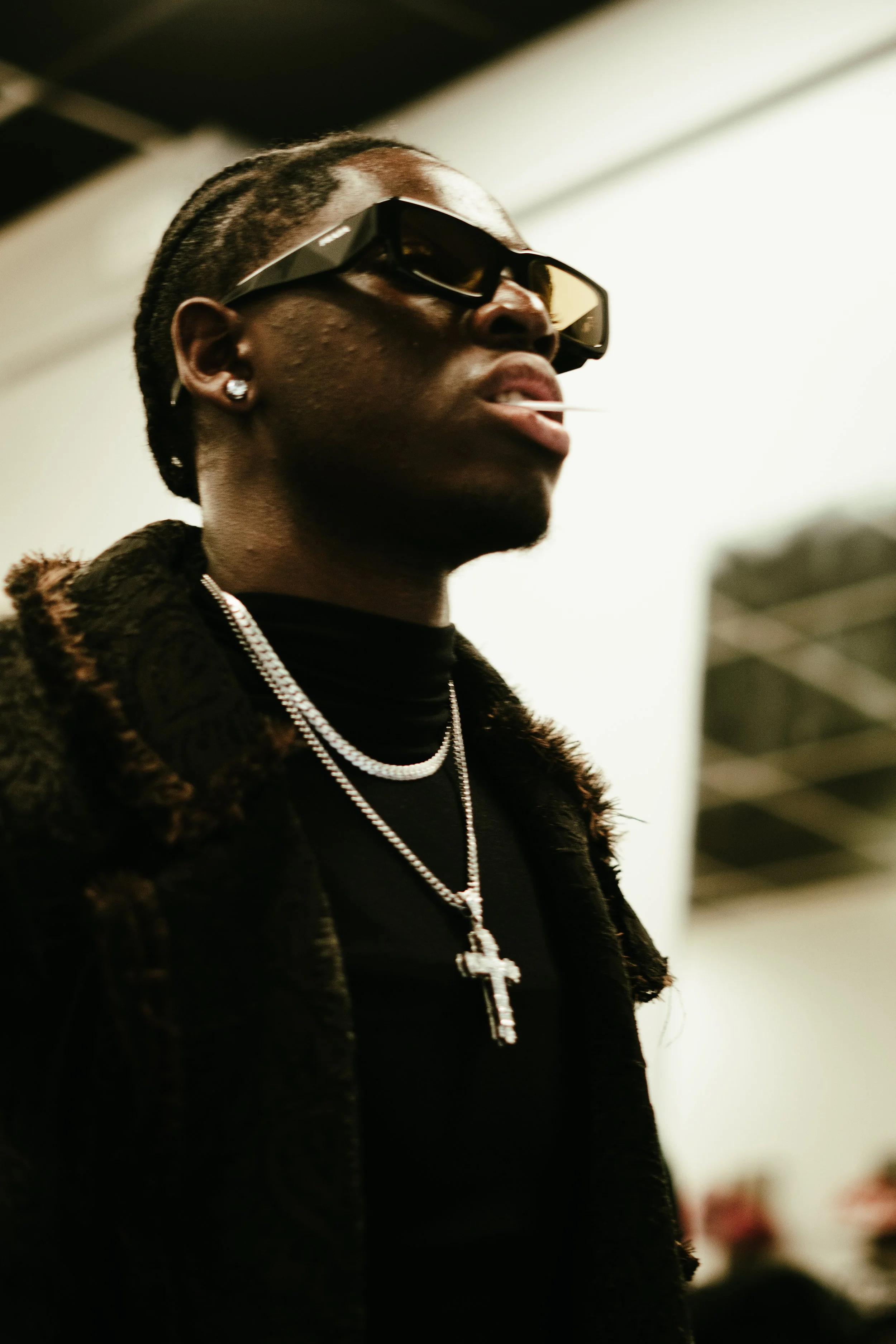 Un homme portant des lunettes de soleil, des bijoux en or, une veste en fourrure, et un cigare dans la bouche dans un intérieur