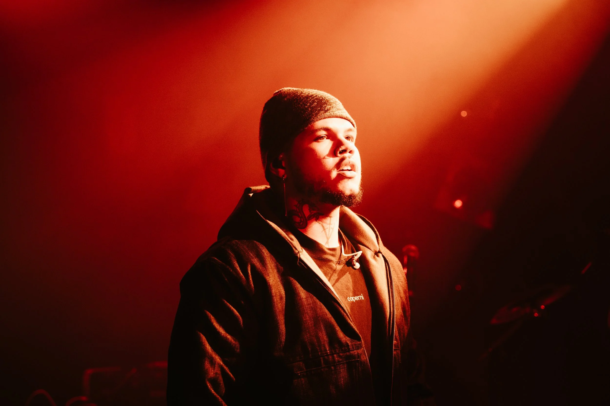 Un homme avec une casquette et une barbe, sous un éclairage rouge, regarde pensivement vers la lumière.