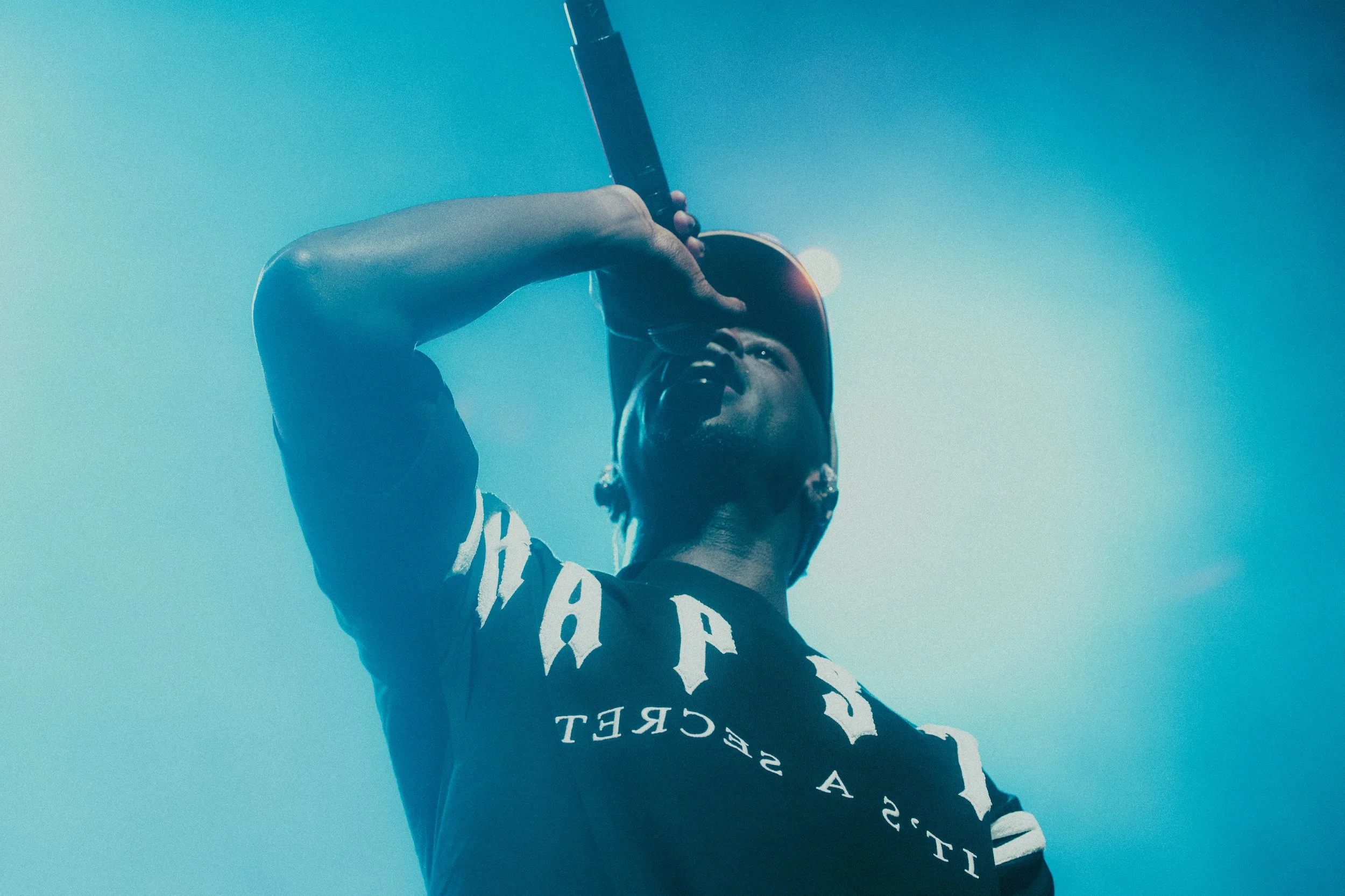 Un homme portant une casquette chante ou parle dans un microphone, vu de dessous, avec un ciel bleu en arrière-plan.