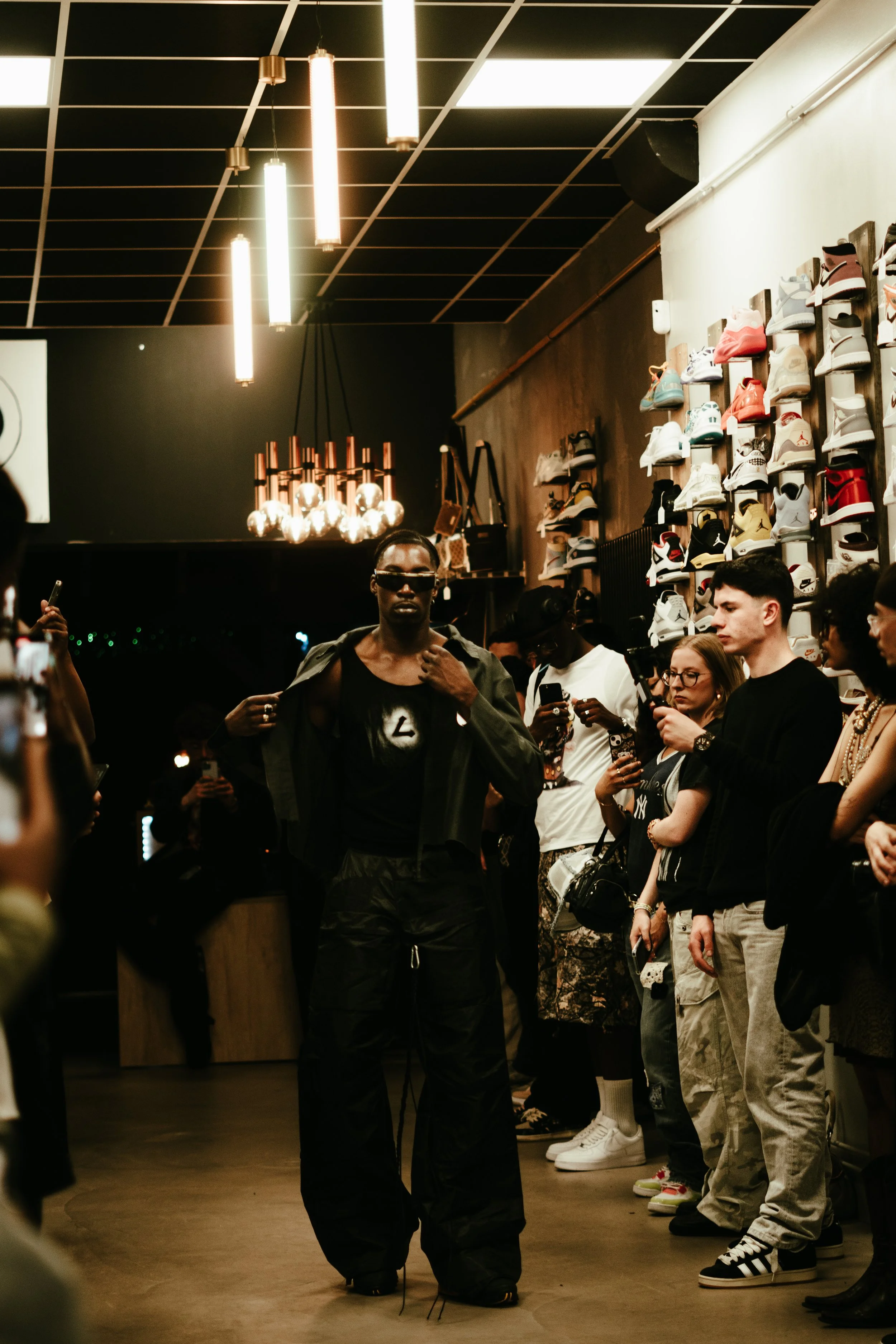 Un homme défile lors d'un défilé de mode dans une boutique avec des chaussures en arrière-plan. Il porte des vêtements noirs et un manteau, et porte des lunettes de soleil.