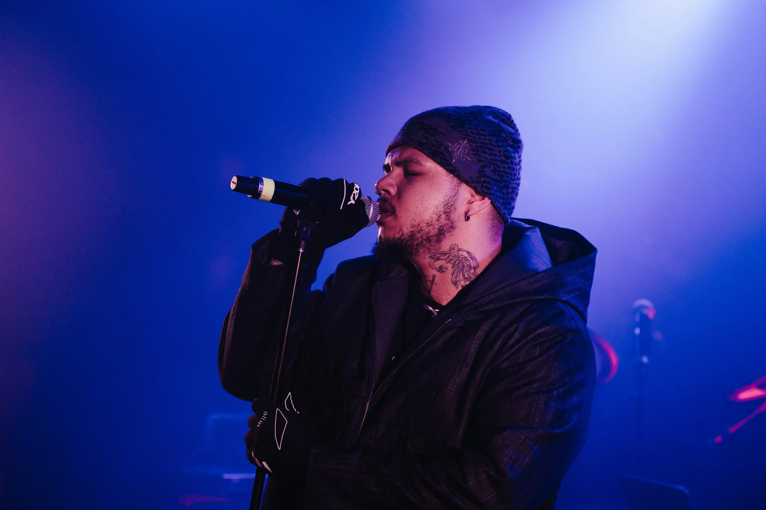 Un homme chante dans un micro, portant un bonnet et des gants noirs, sous un éclairage violet et bleu, avec un tatouage visible sur le cou.