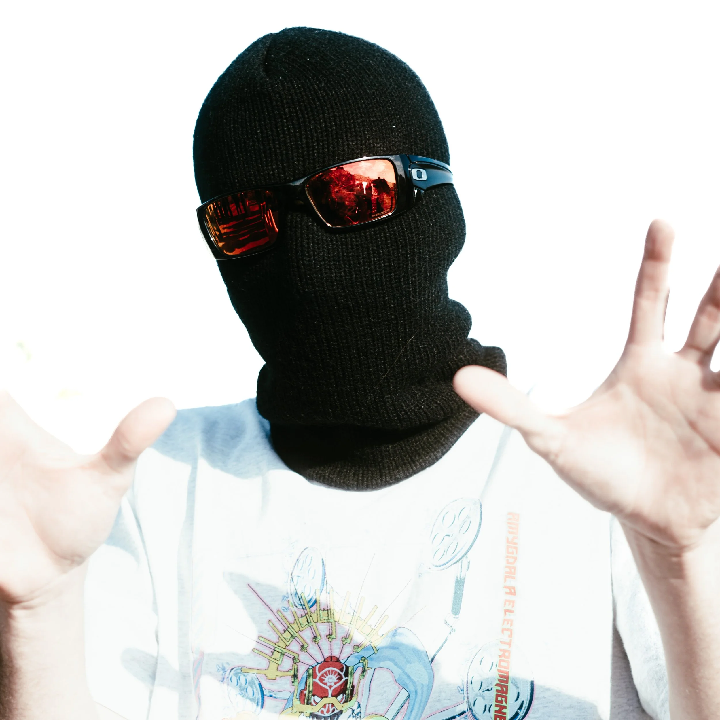 Person portant un masque noir, des lunettes de soleil noires réfléchissant la scène, et un t-shirt blanc avec un motif complexe, faisant un geste de la main. arrière-plan blanc.
