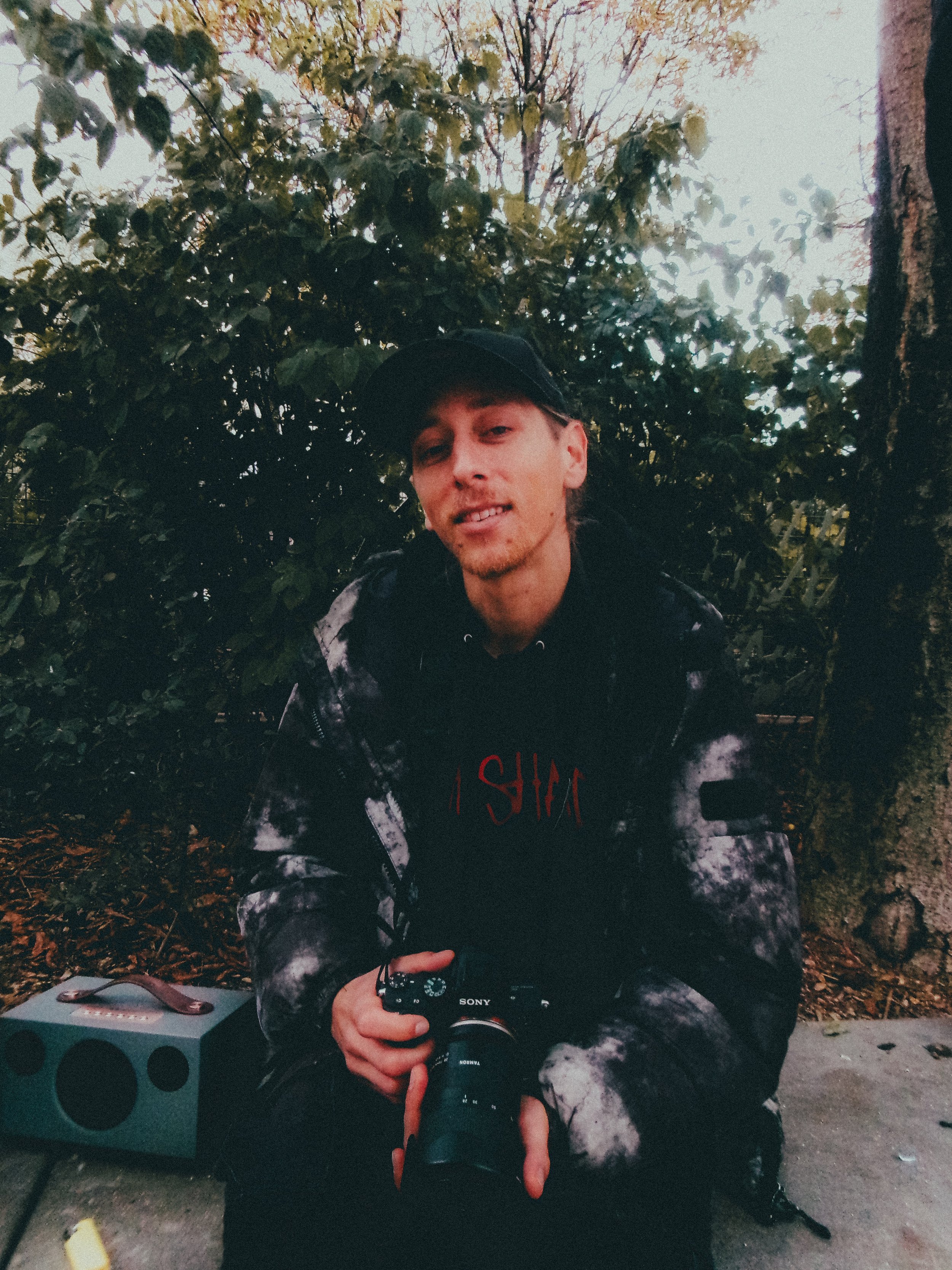 Jeune homme assis dehors dans la nature, tenant un appareil photo Sony, avec un fond de feuillage vert et un arbre à droite.