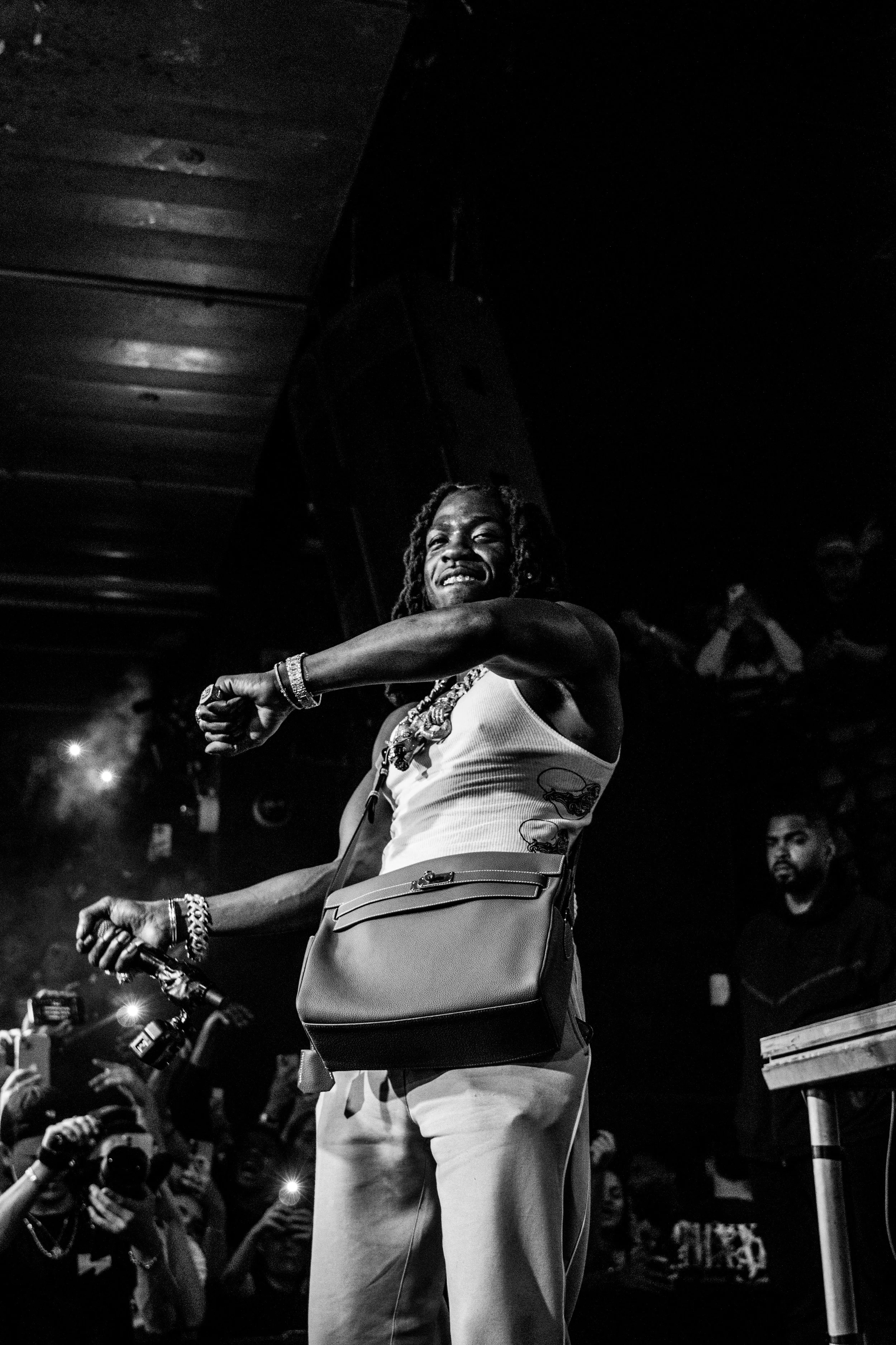 Une femme souriante avec des dreadlocks, portant un débardeur blanc avec des motifs, un sac à main et des bracelets, lors d'une performance ou d'un événement, entourée d'une foule de photographes et spectateurs. L'image est en noir et blanc.