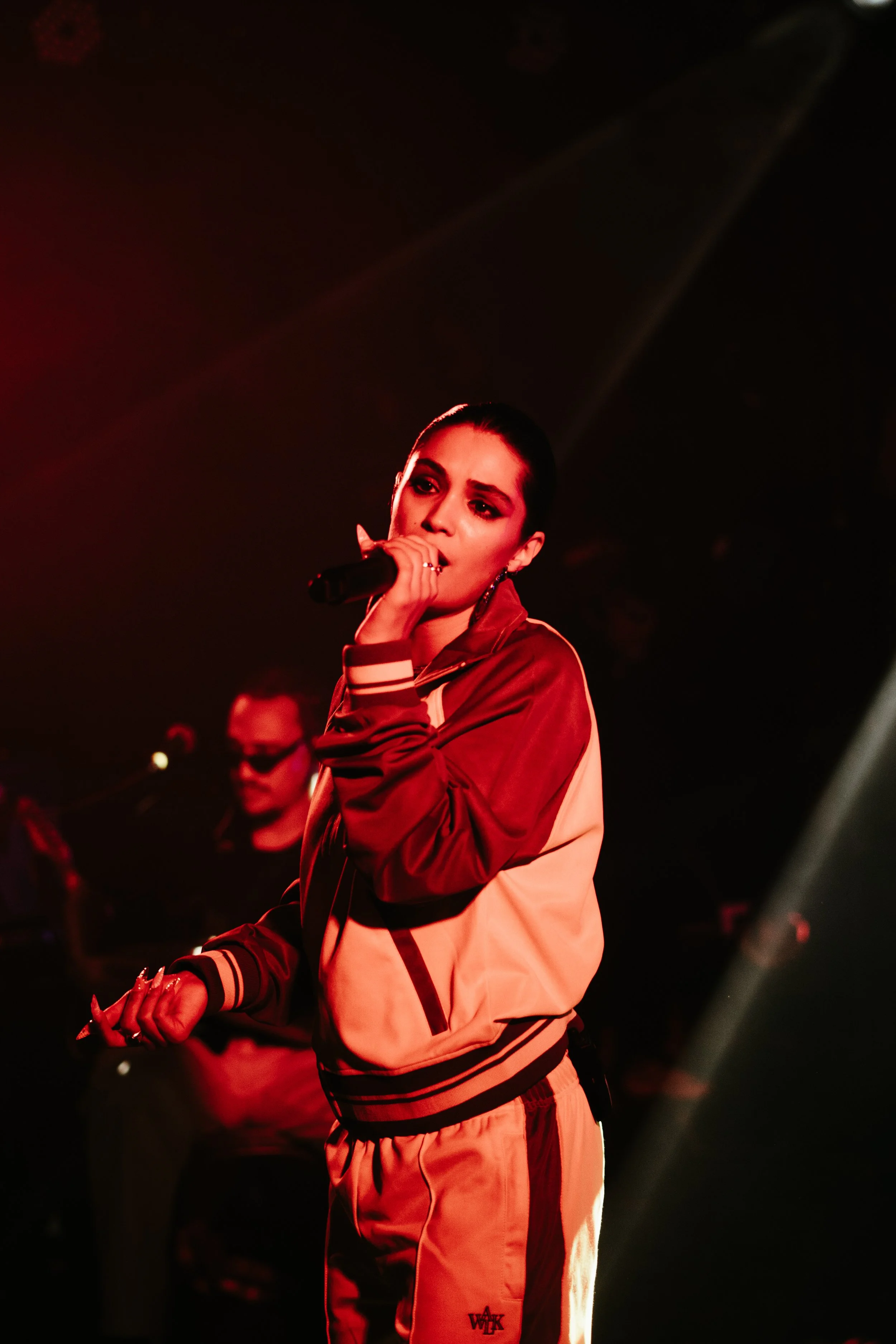 Une femme chante en tenant un microphone sur scène, avec un éclairage rouge foncé. Elle porte un survêtement avec des couleurs rouge et beige et a une expression concentrée.