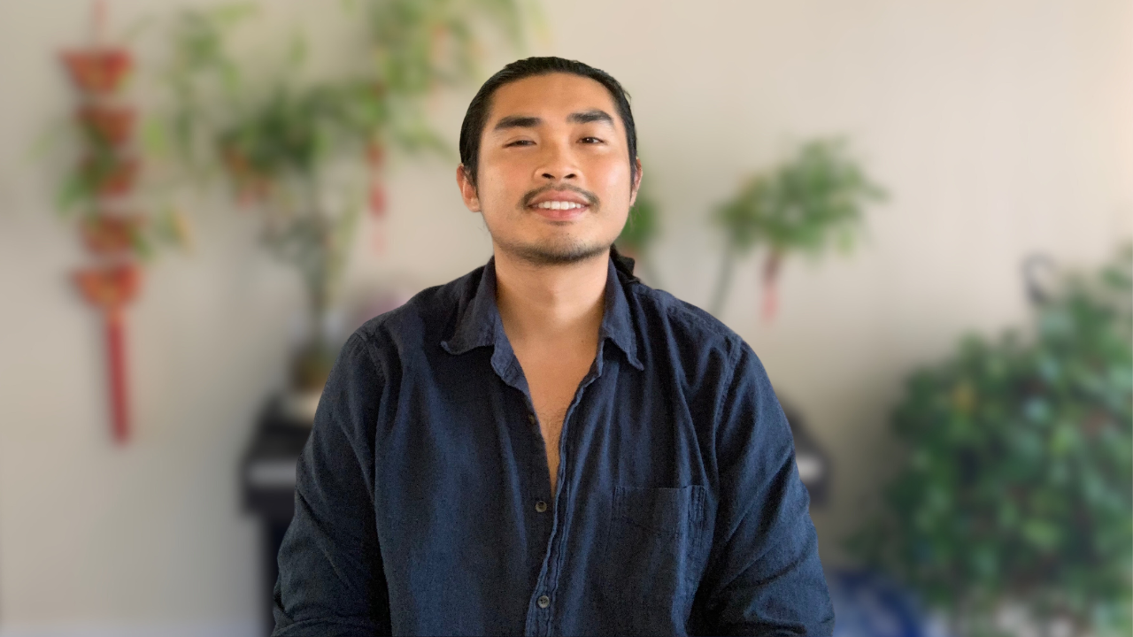 A young man with dark hair and facial hair sitting in front of a blurred background with plants and a wall