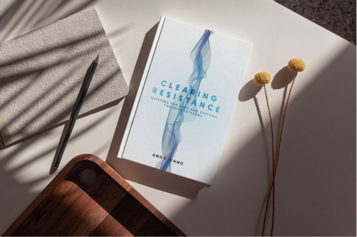 A book titled "Clearing Resistance" by Andy Wang, a notebook, a pen, a wooden tray, and two yellow billy ball flowers on a white surface with shadows.