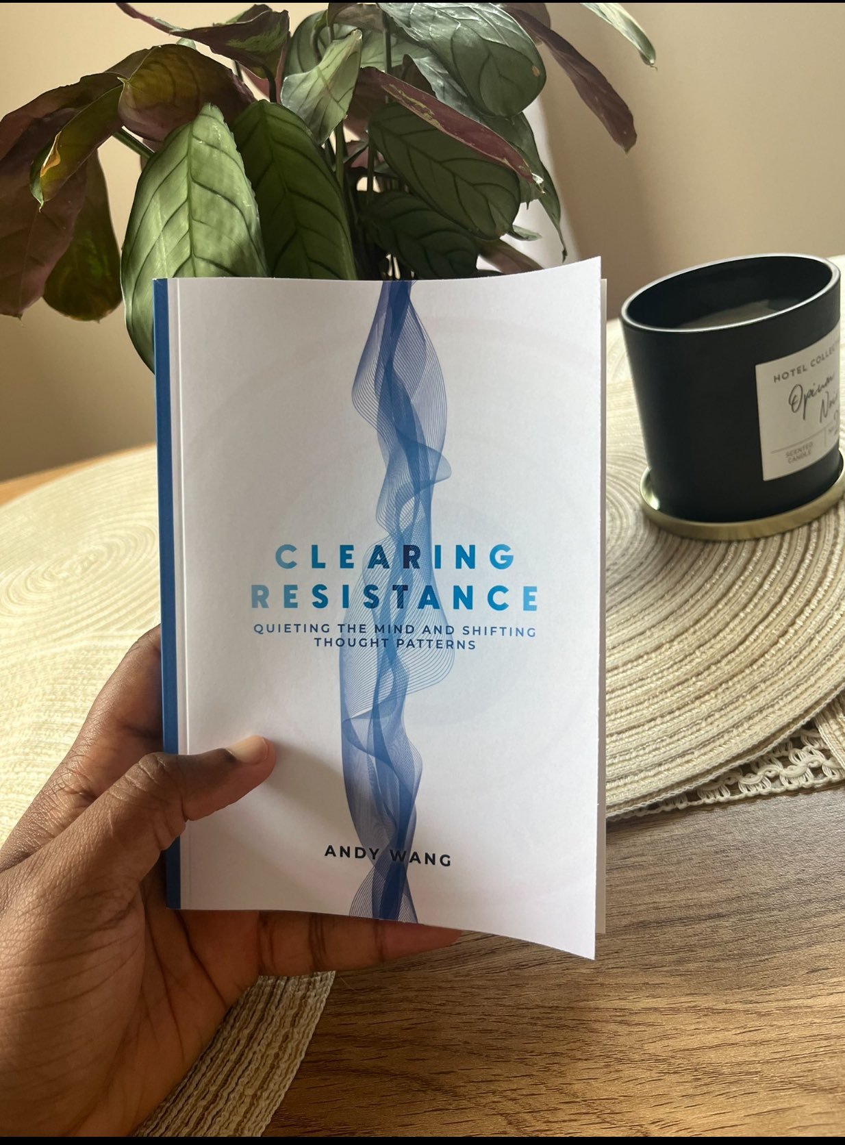 A person holding a book titled 'Clearing Resistance: Quieting the Mind and Shifting Thought Patterns' by Andy Wang in front of a potted plant and a black candle holder with a 'Hotel Collection' candle on a round woven placemat on a wooden table.
