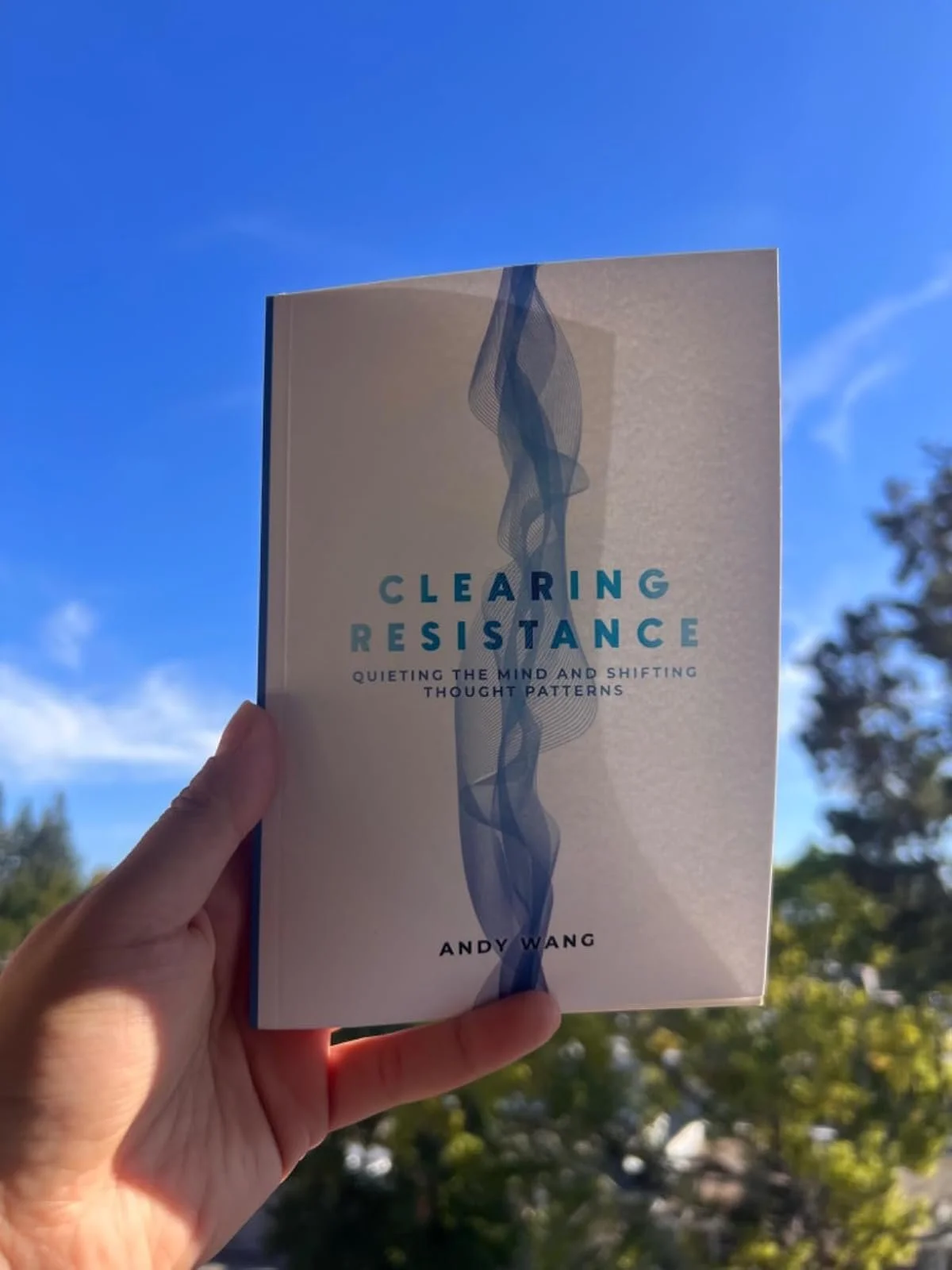 A person holding a book titled 'Clearing Resistance: Quieting The Mind And Shifting Thought Patterns' by Andy Wang against a blue sky with trees in the background.