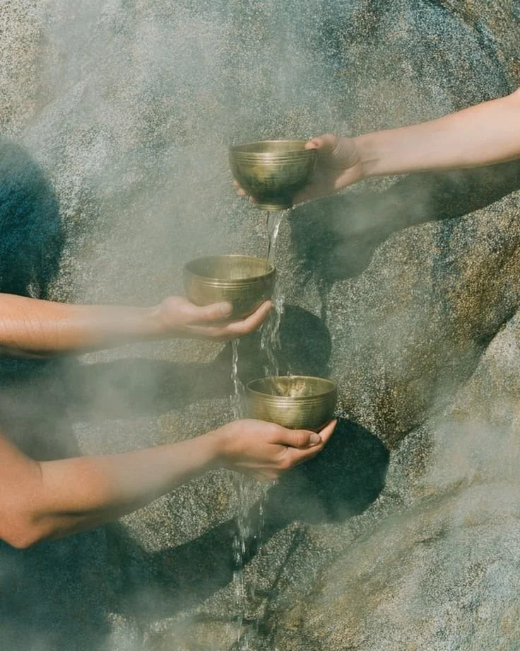 Three hands holding brass bowls with water flowing between them, against a textured rock wall as a cleansing ritual for clearing resistance.