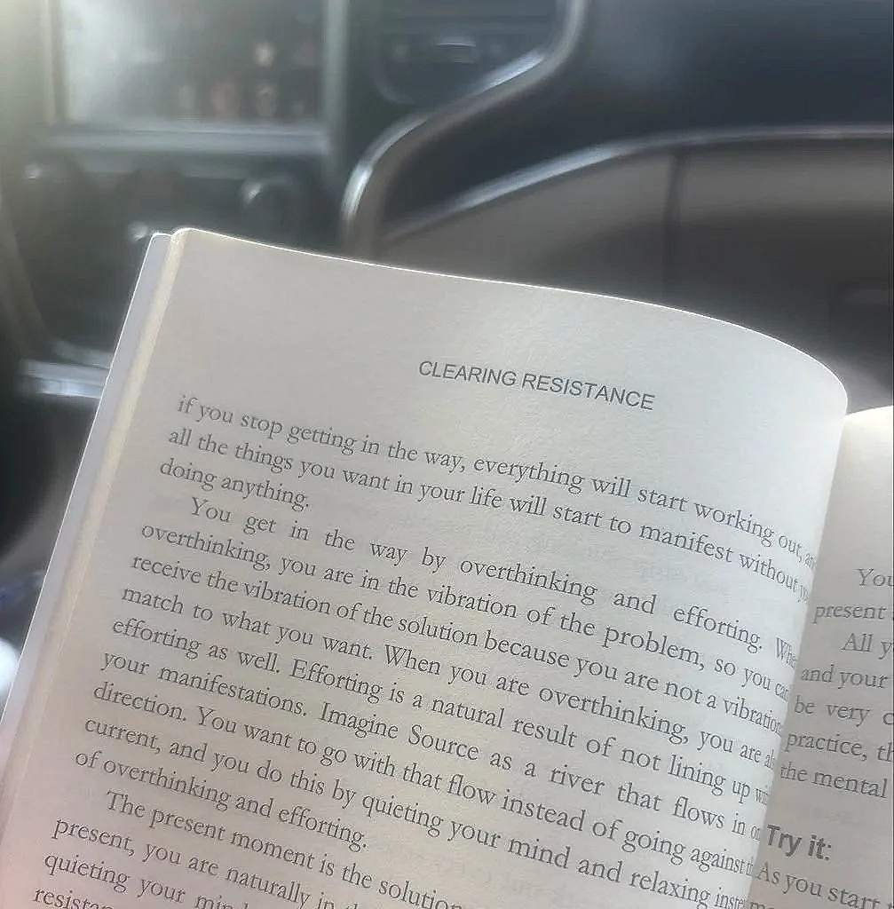 Open book with text on a seat or dashboard of a vehicle, titled "Clearing Resistance" discussing overcoming overthinking and vibration to manifest desires.