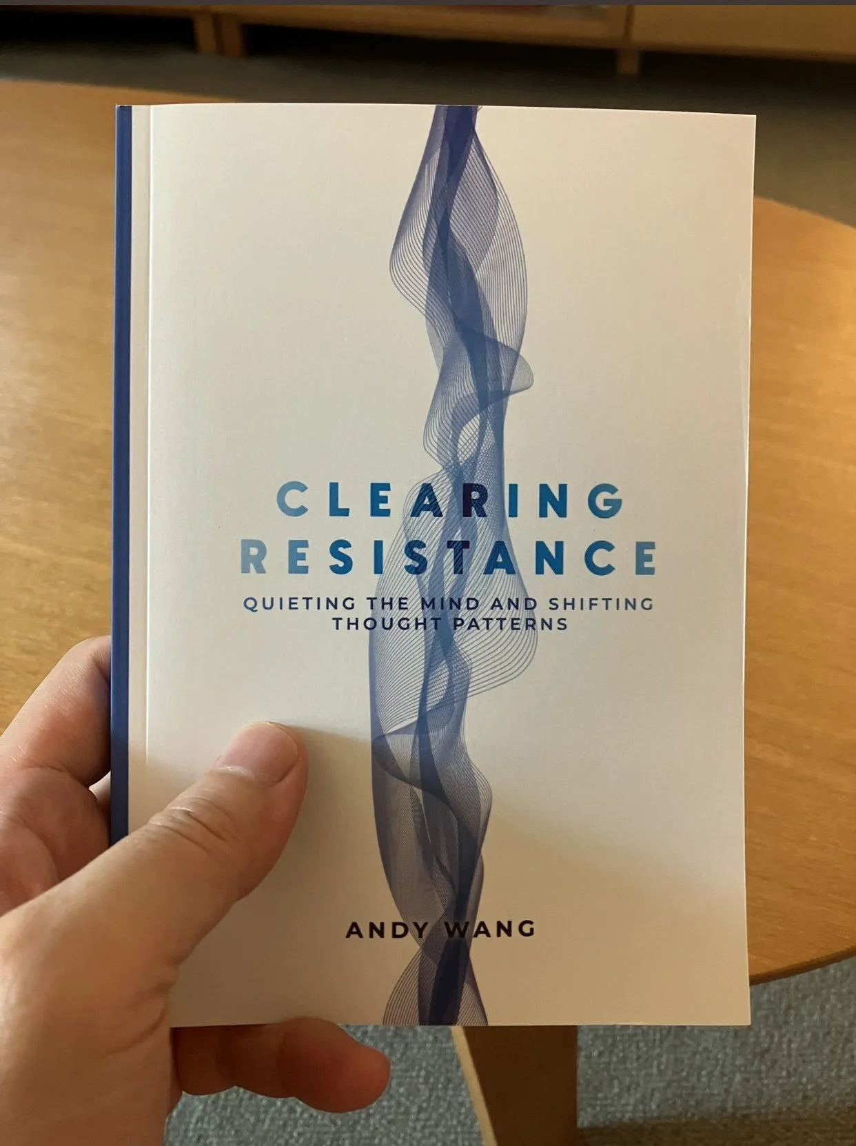 A person holding a book titled "Clearing Resistance" by Andy Wang. The cover has a blue and gray abstract wavy design and the subtitle "Quieting the Mind and Shifting Thought Patterns."
