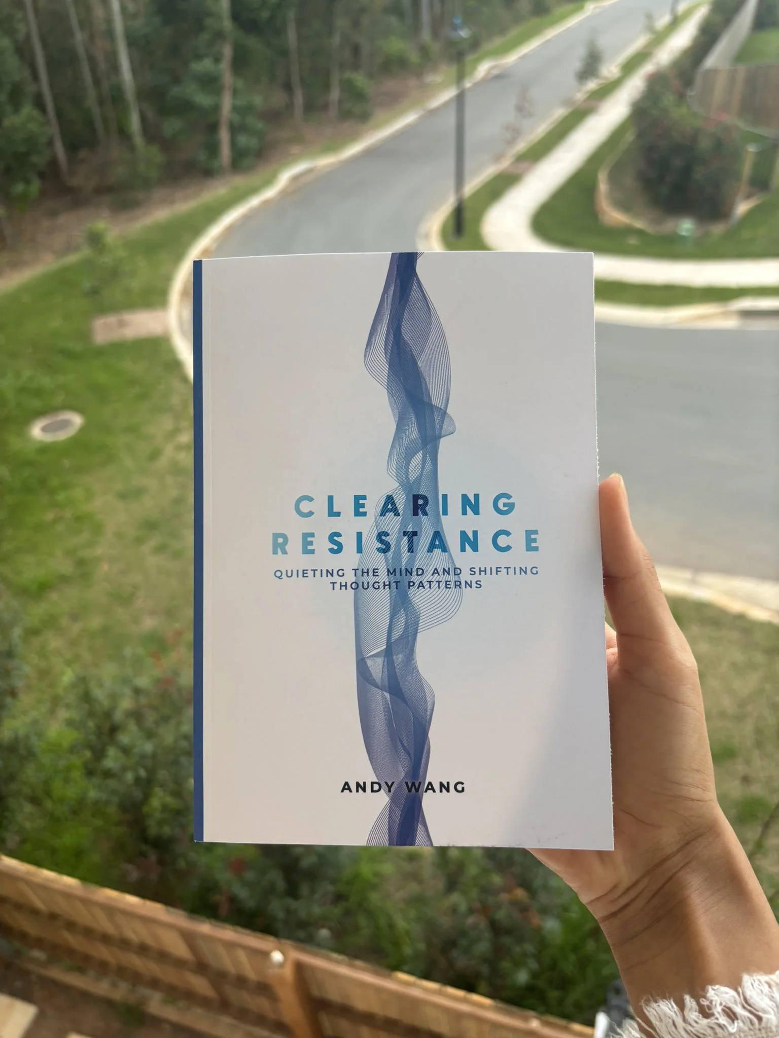 A person holding a book titled 'Clearing Resistance' by Andy Wang in front of an outdoor scene with a curved road, green grass, trees, and a sidewalk.