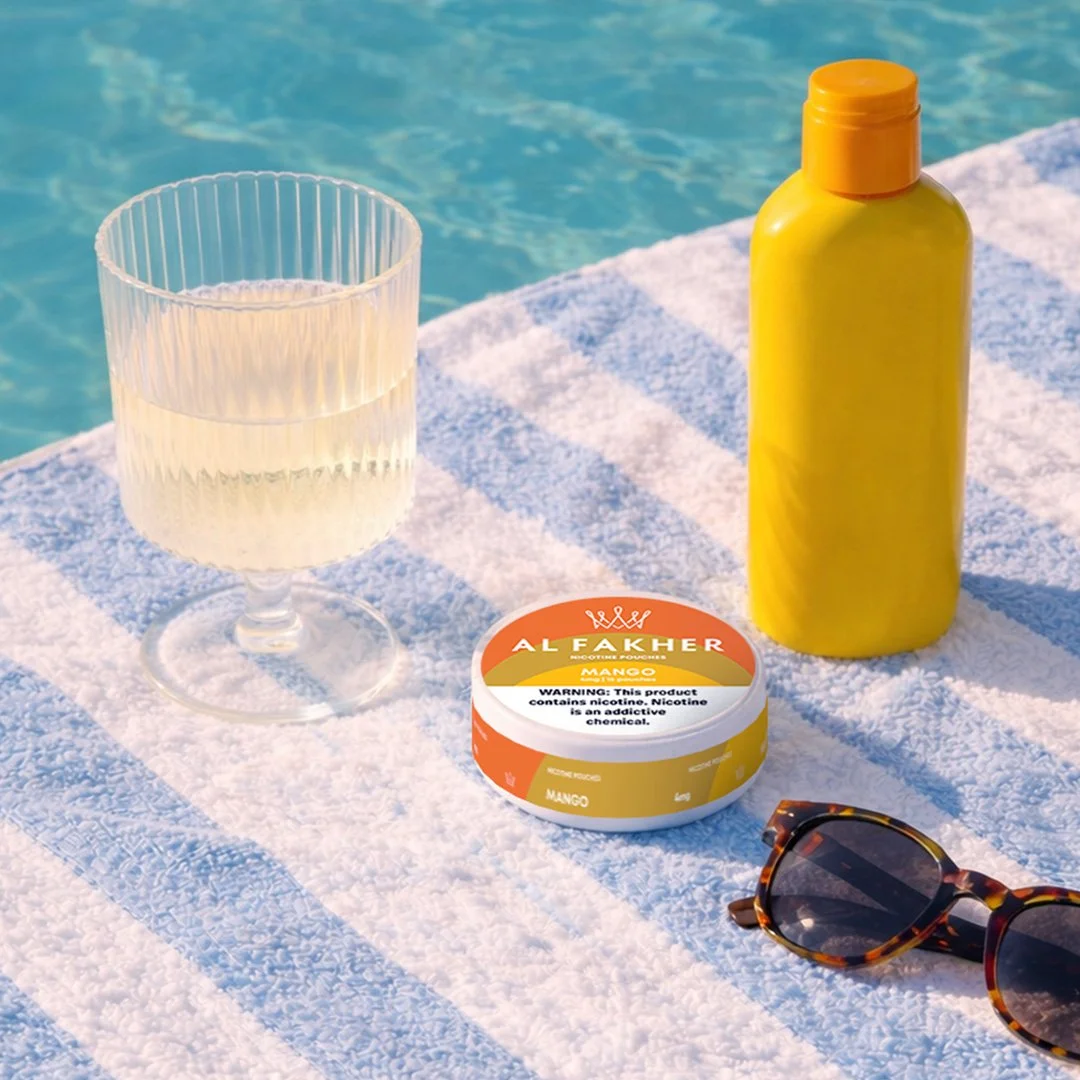 Al Fakher Mango Nicotine Pouches can on a poolside towel with sunglasses and a yellow bottle.