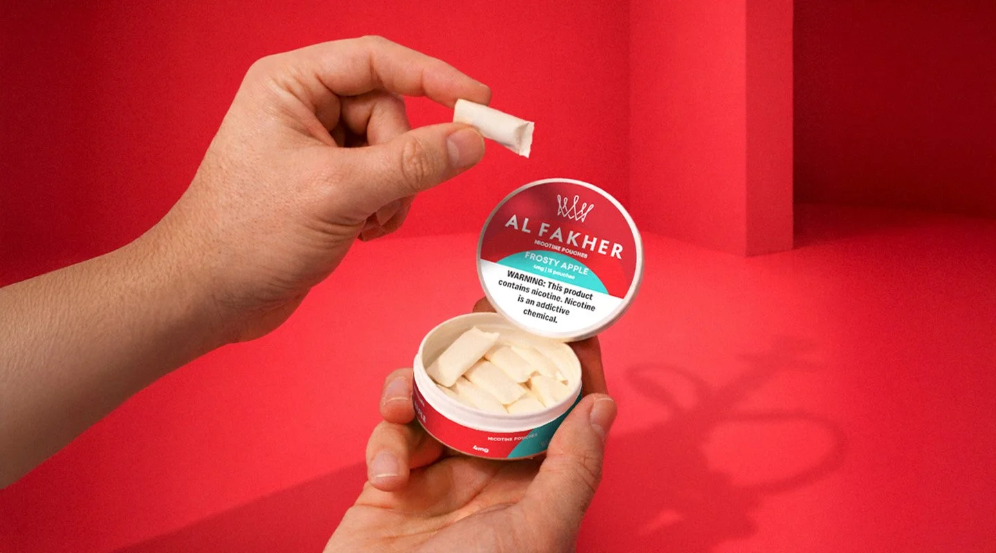 Hand picking up an Al Fakher Frosty Apple Nicotine Pouch from an open can on a red background.