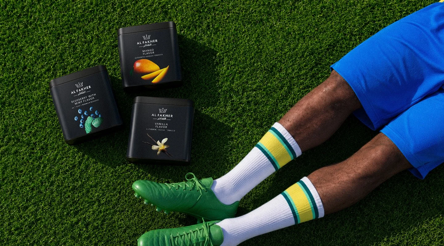 Al Fakher Mango, Blueberry with Mint and Vanilla tubs arranged on a football pitch beside a player in Brazil-inspired kit.