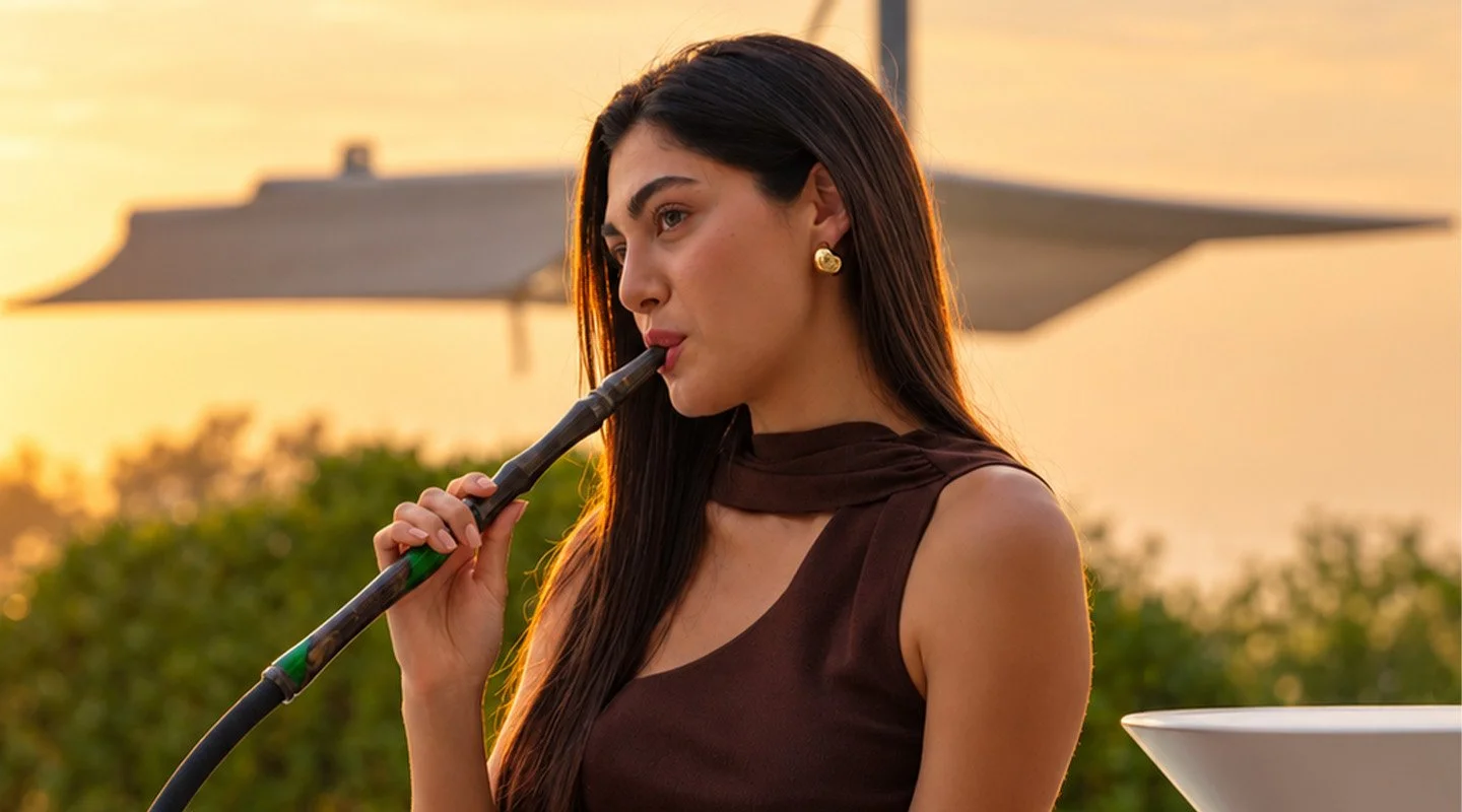 Woman using a shisha pipe outdoors at sunset, illustrating the relaxed atmosphere and social traditions associated with modern shisha culture.