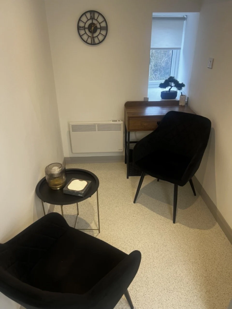 A clear clinic room with two black armchairs, a side table with a candle and tissue box, a wooden desk with a small bonsai tree, a window showing outdoor trees. The space feels calm, clean and inviting.