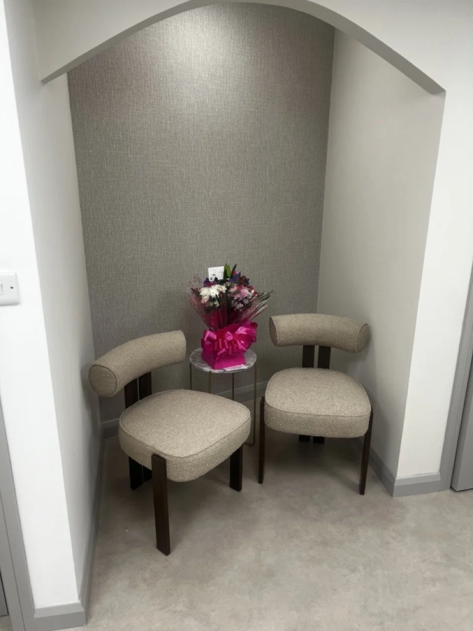 Two beige upholstered chairs with dark wooden legs are positioned under an archway, with a small round table between them holding a bouquet of flowers wrapped in pink paper.