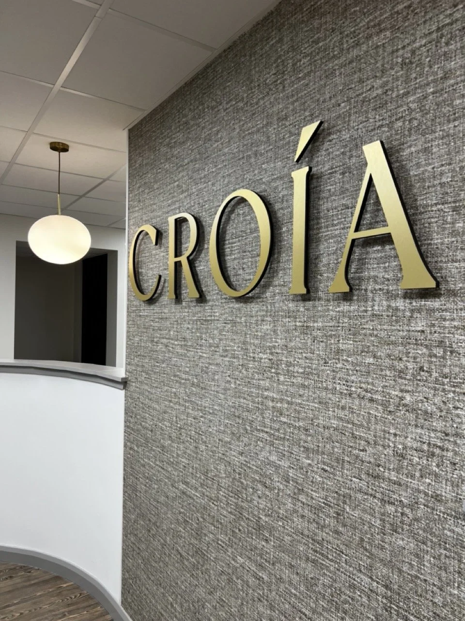 Bright and airy clinic space in Kilmarnock Ayrshire with textured grey wall displaying the word 'CROIÁ' in large gold letters, a white curved reception desk, and a round ceiling light.