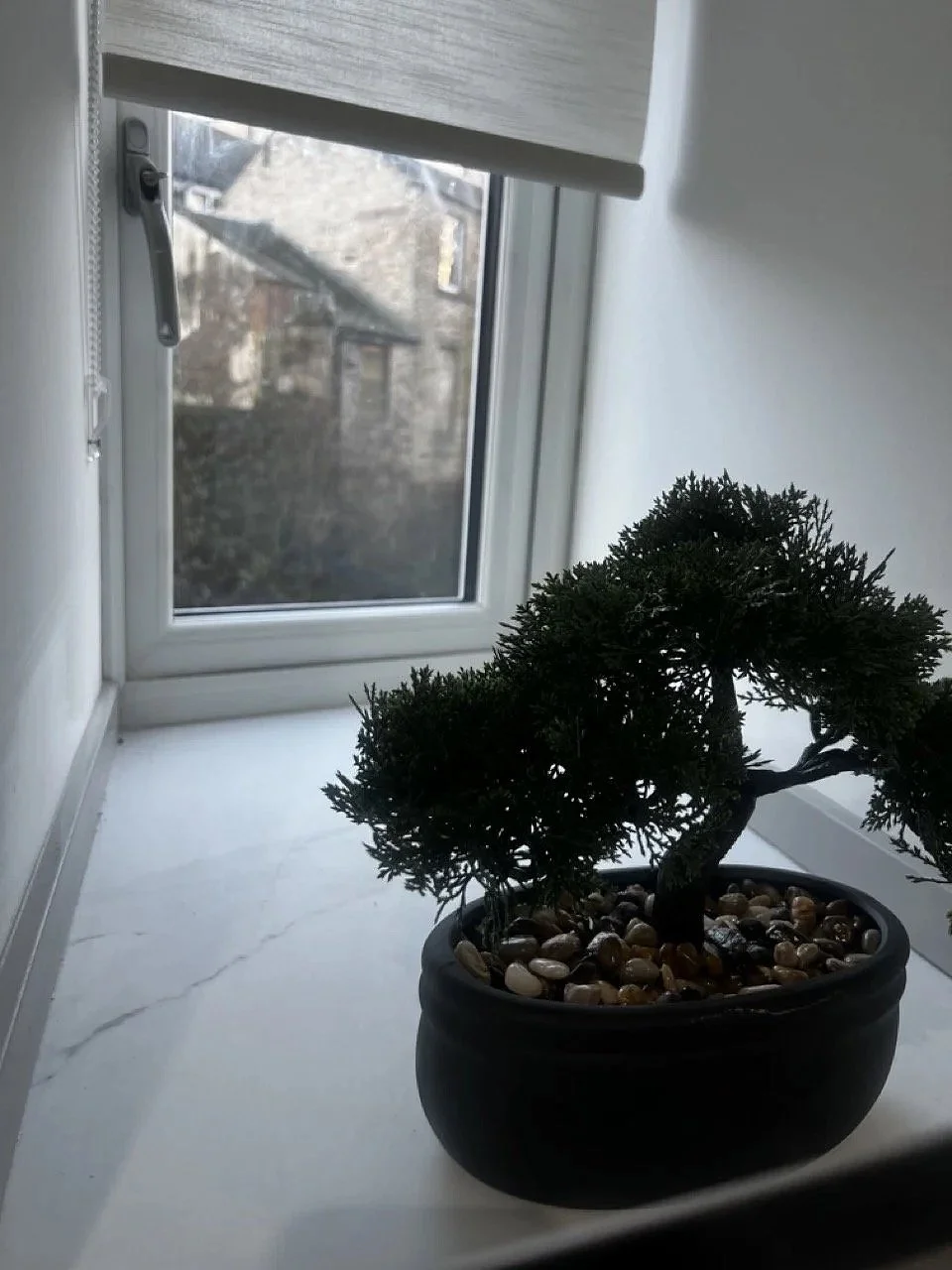 Small potted bonsai tree with rocks on soil, placed on white window ledge near a partially open window with a white frame and a view of trees outside.