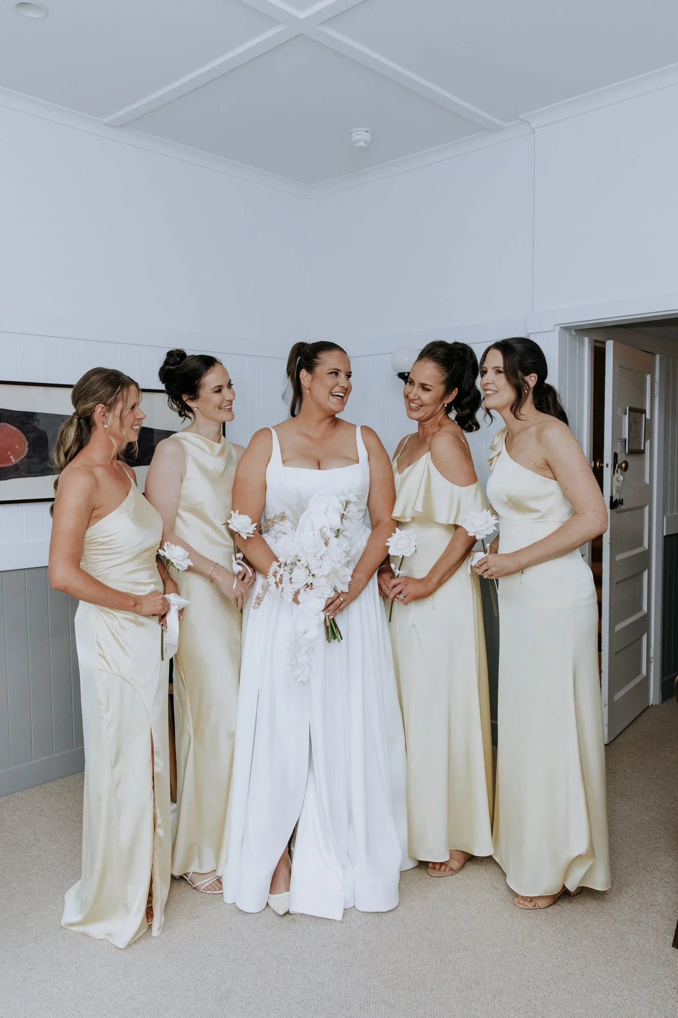 A bride in a white wedding dress holding a bouquet of white flowers stands with her bridesmaids, who are wearing matching pale yellow satin dresses, inside a light-colored room.