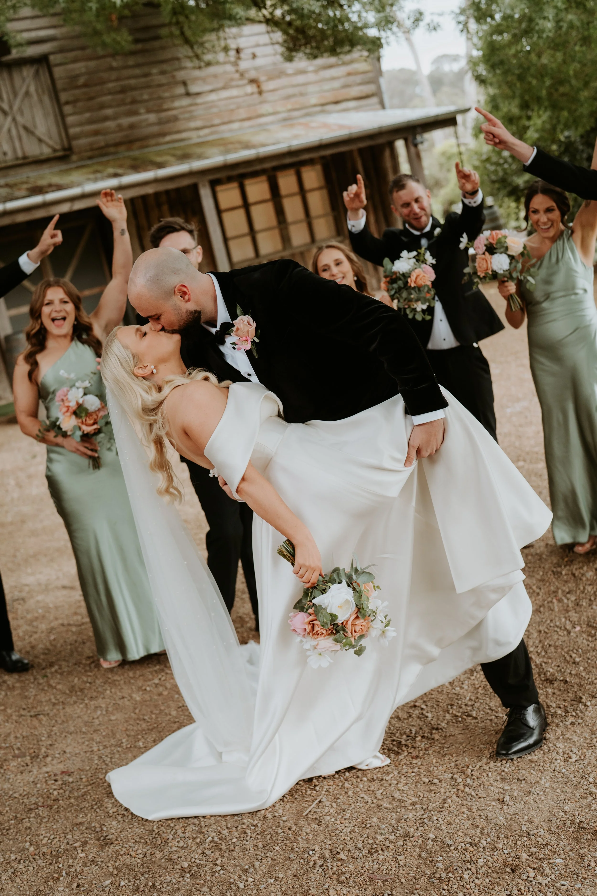 A newlywed couple kisses during a wedding celebration, with the groom dipping the bride, surrounded by bridesmaids and groomsmen holding bouquets and celebrating outdoors.