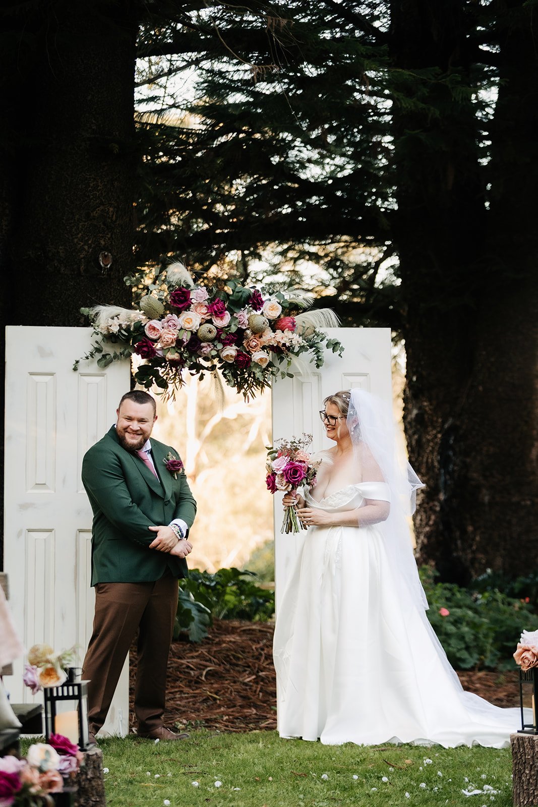 A newlywed couple at their wedding ceremony outdoors, standing in front of a white decorative backdrop with pink, purple, and white floral arrangements, with large trees in the background.