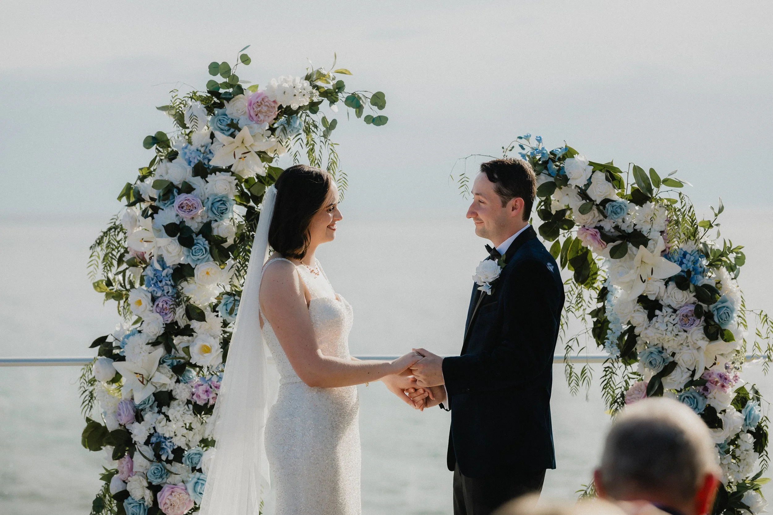 A bride and groom holding hands during their outdoor wedding ceremony, standing in front of a floral arch with white, pink, and blue flowers at a beach.