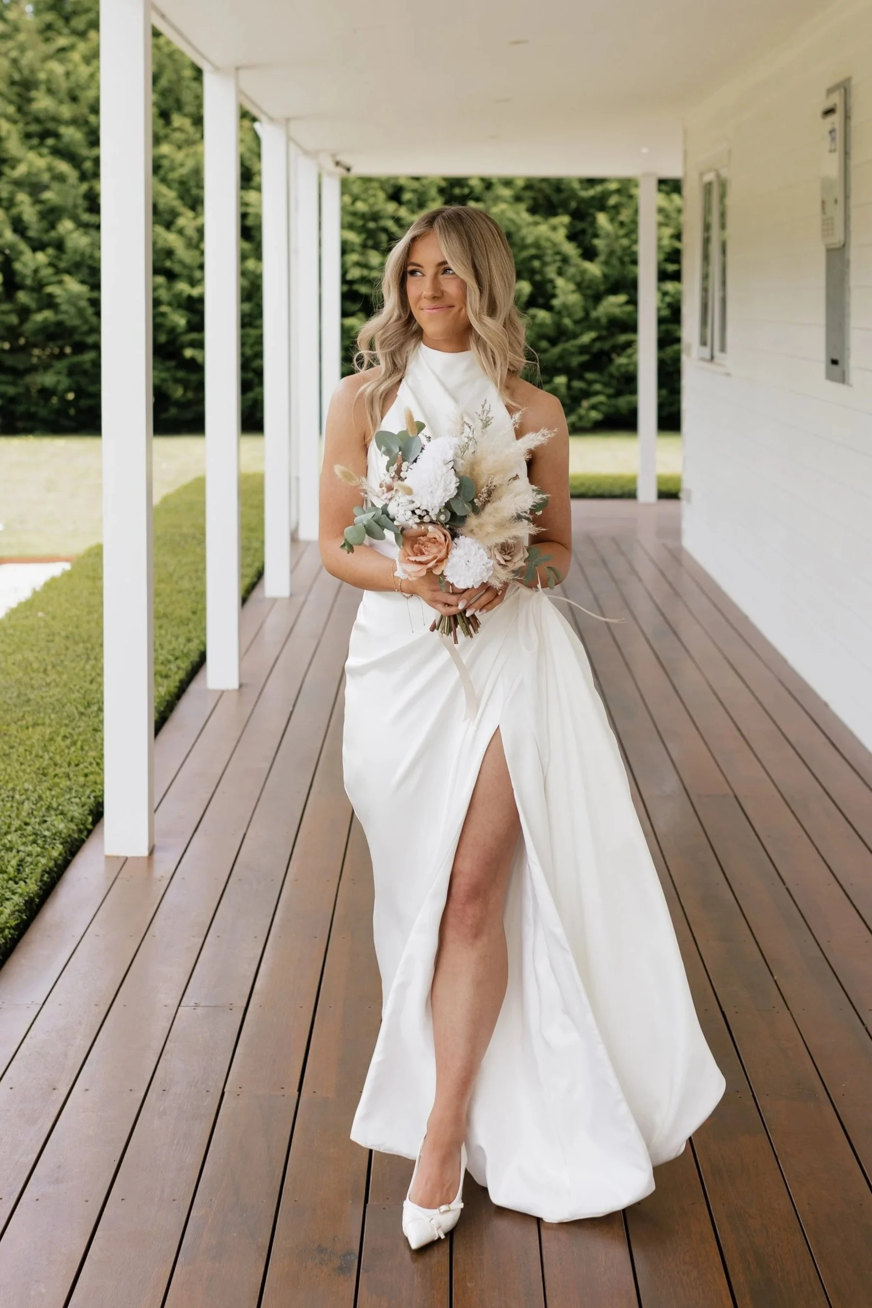A bride in a white wedding dress walking on a wooden porch while holding a bouquet of flowers, with a background of green trees.