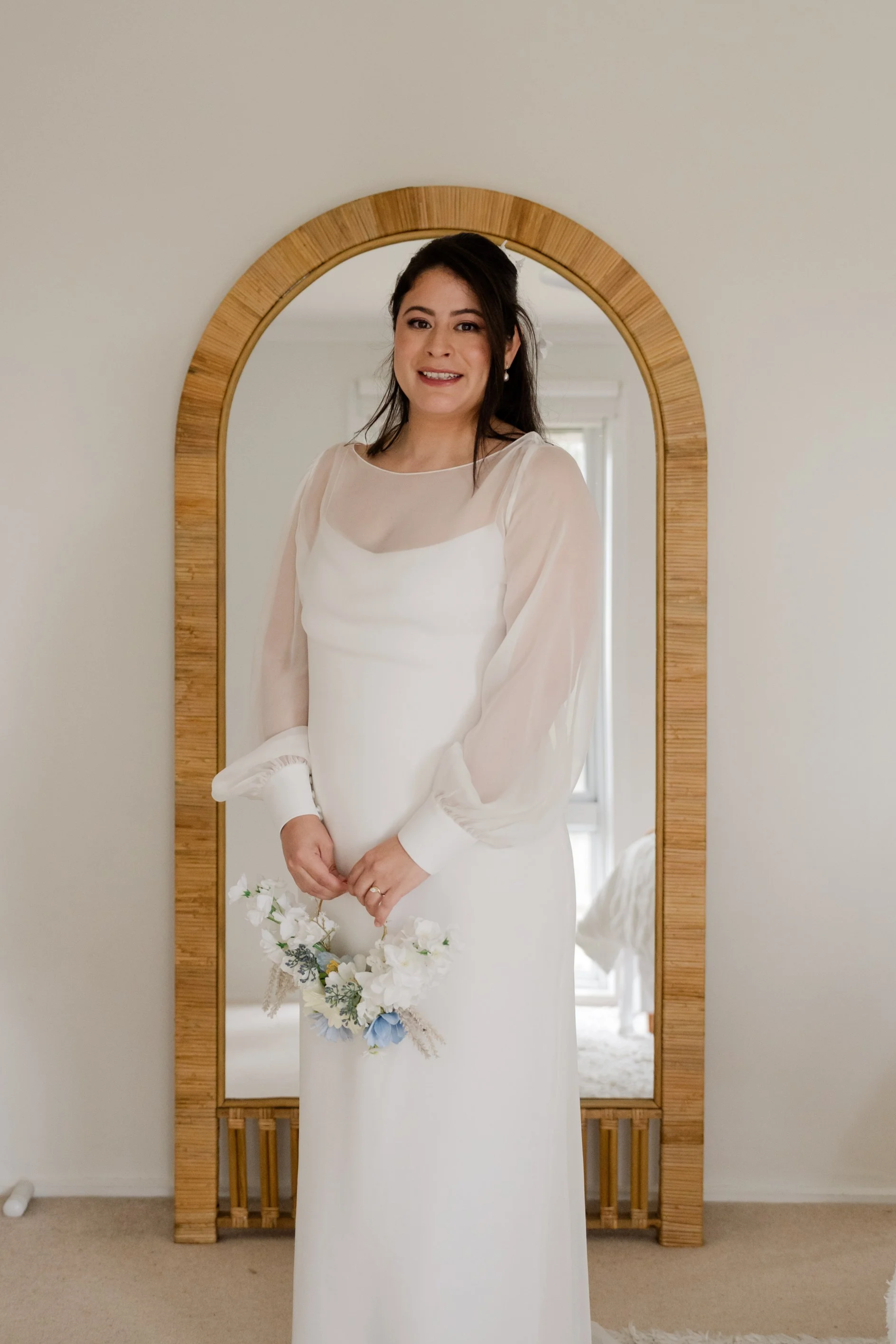 A woman in a white dress standing in front of a full-length wooden framed mirror, holding a flower bouquet, smiling.
