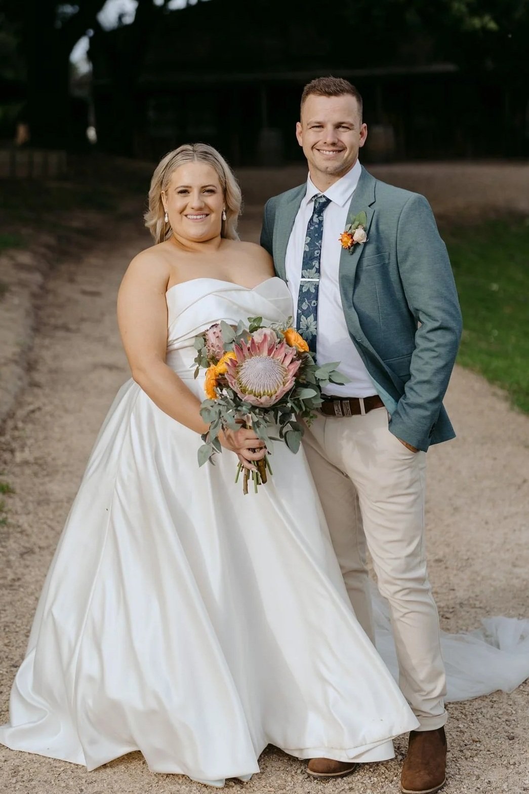 A bride and groom standing outdoors on a dirt path, smiling at the camera. The bride wears a strapless white wedding gown and holds a bouquet with pink protea, orange flowers, and greenery. The groom wears a teal blazer, floral tie, white shirt, beig