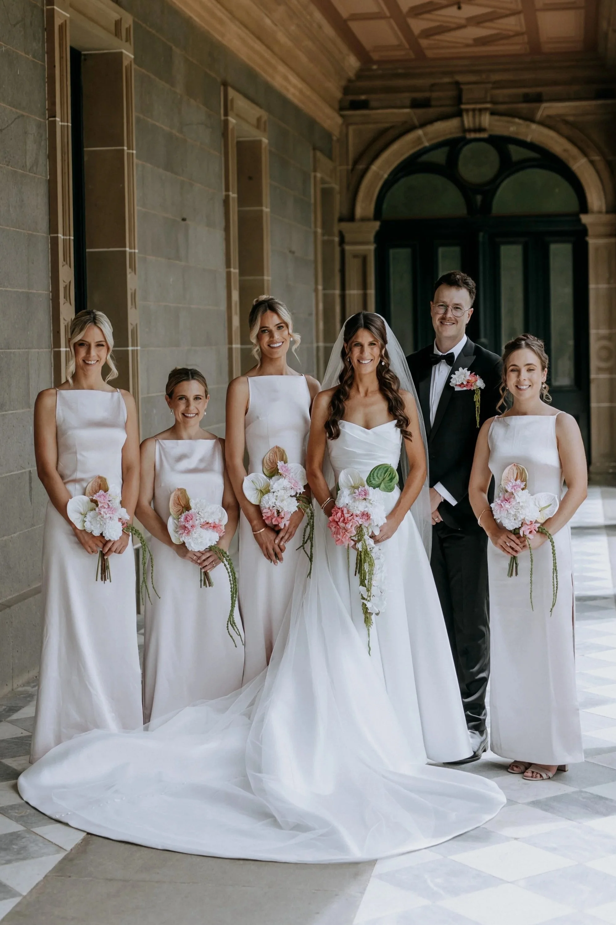Wedding party standing together indoors, with the bride and groom in the center. The bride wears a white gown with a long train, and the groom in a black tuxedo with a bow tie. Four bridesmaids in matching white dresses hold bouquets of pink and whit