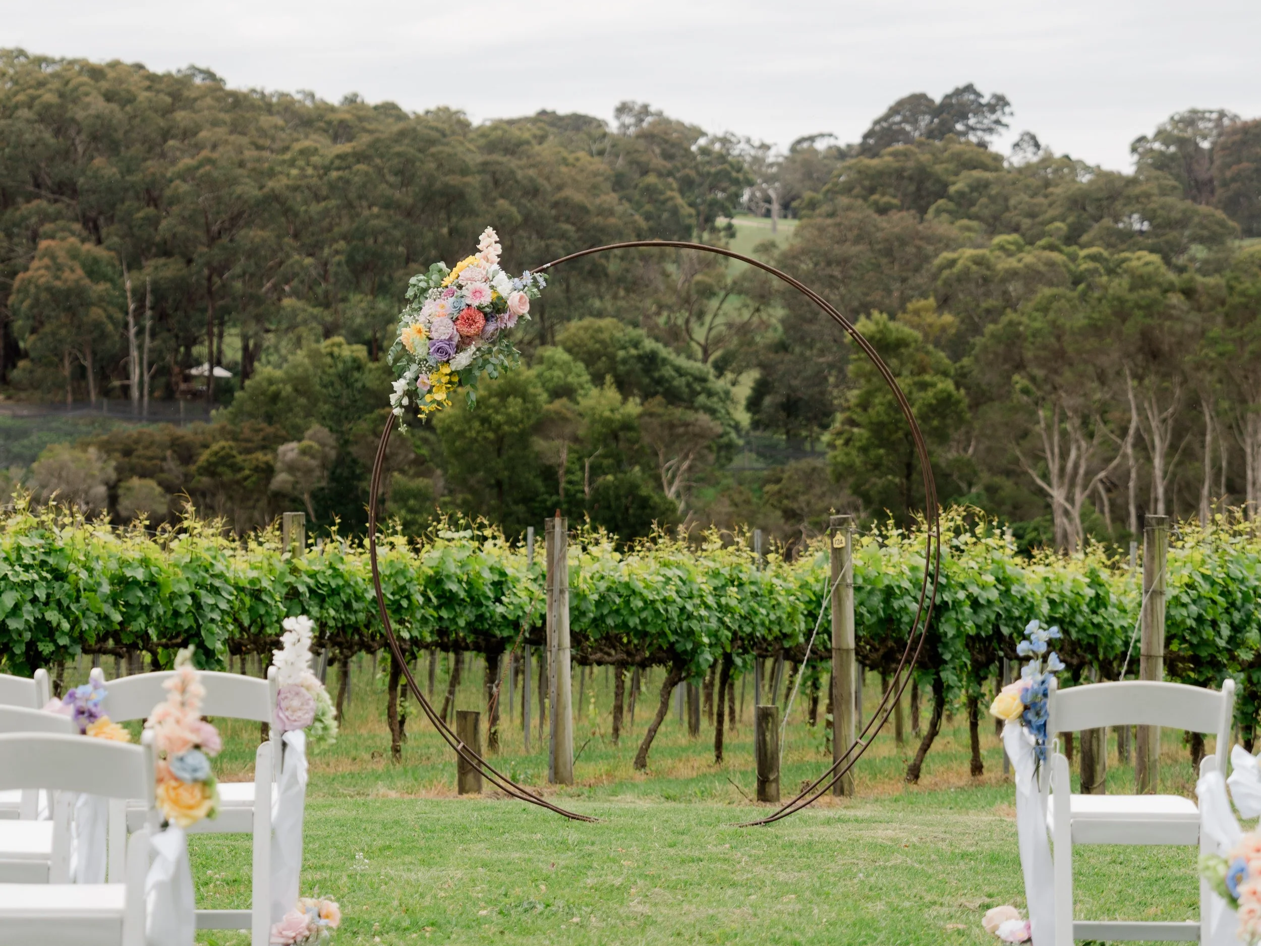 Outdoor wedding setup with white chairs decorated with flowers, a large circular floral arch, set in a vineyard with green vines and trees in the background.