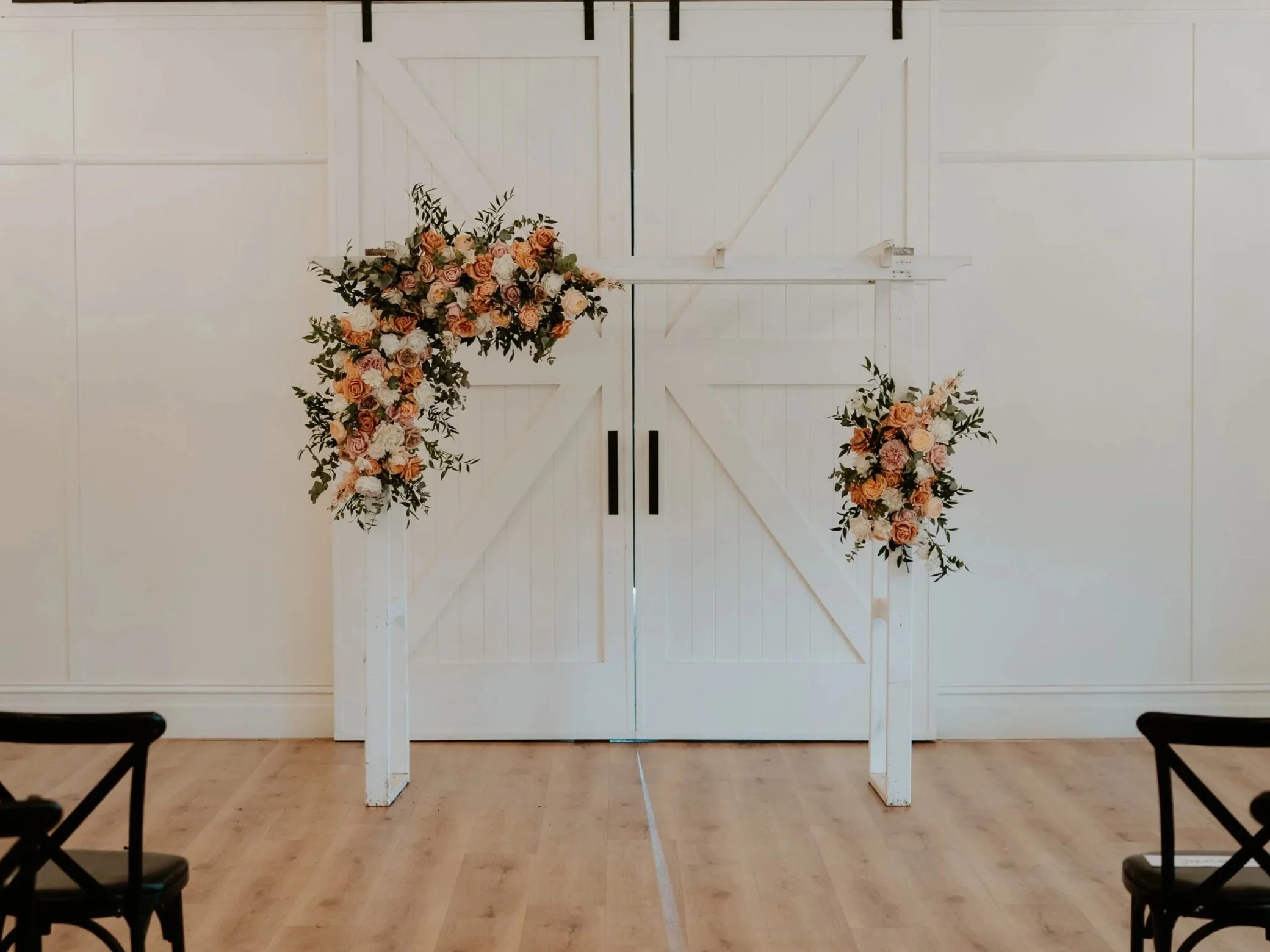 White barn door with floral arrangements on both sides, set up for a wedding ceremony in an indoor venue with light wood flooring and white walls.
