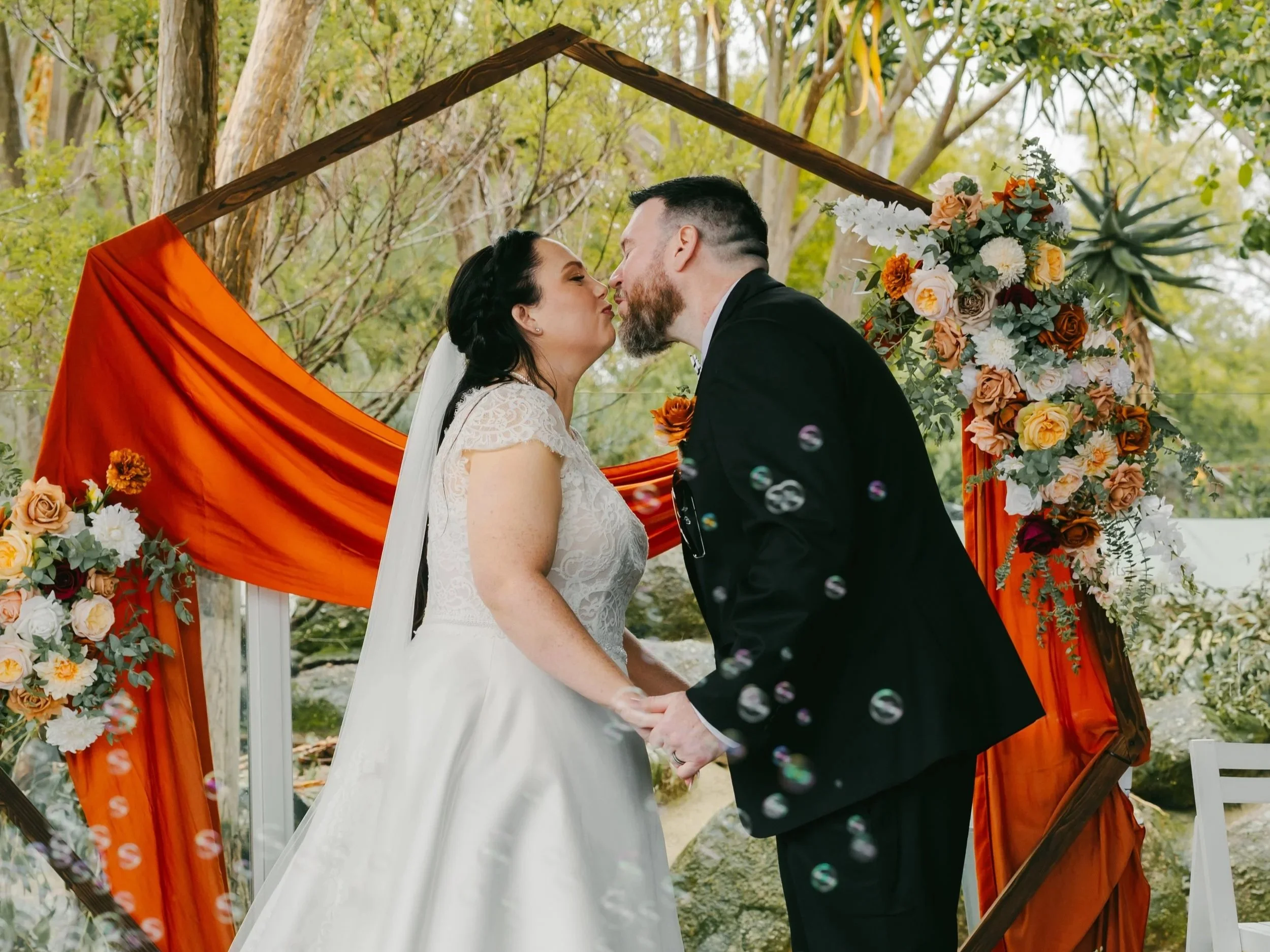 A bride and groom sharing a kiss during their outdoor wedding ceremony, standing in front of a wooden arch decorated with orange fabric and floral arrangements, with trees in the background.