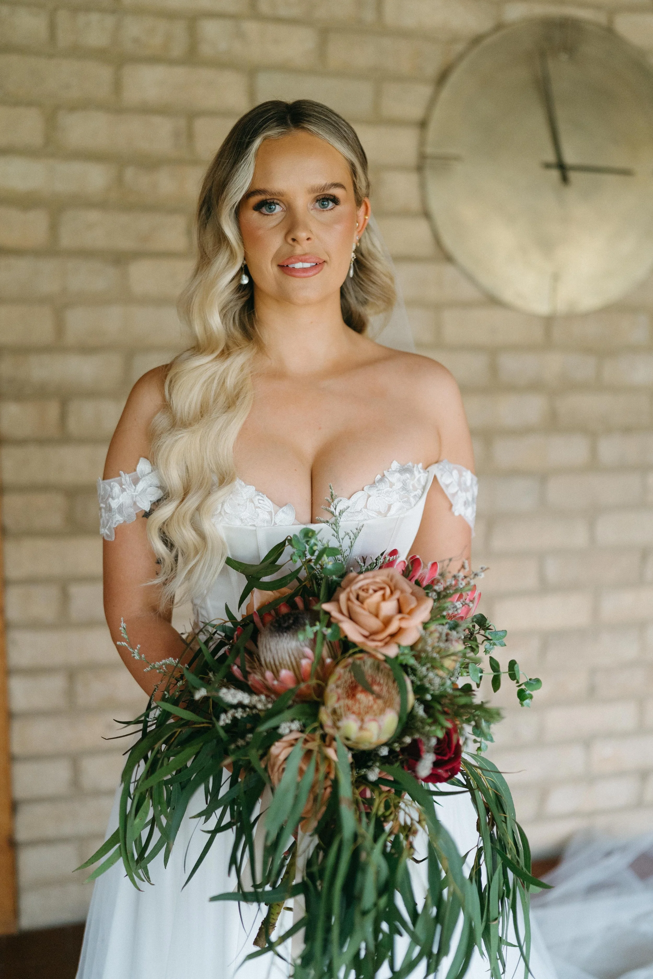 A bride with long, wavy blonde hair in a white wedding dress holding a large bouquet of flowers, standing indoors in front of a brick wall with a round clock.