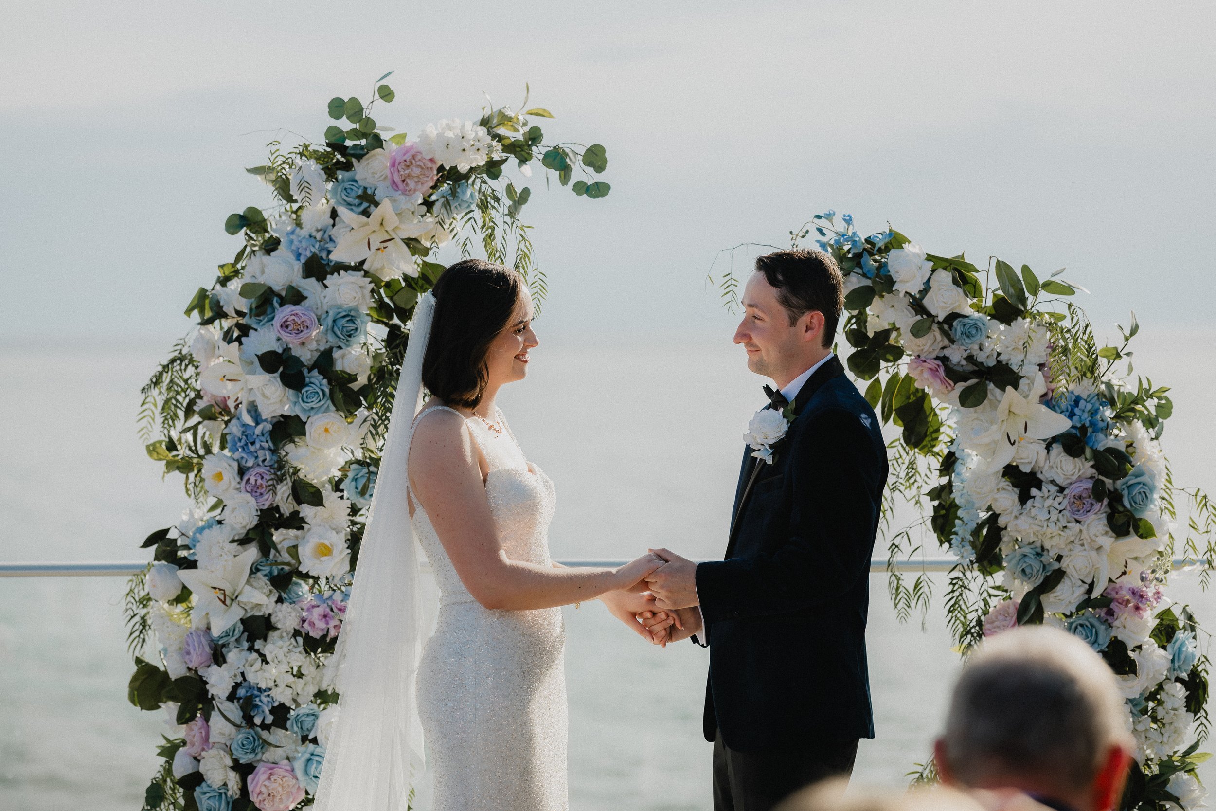 A bride and groom holding hands during their wedding ceremony, standing beneath a floral arch on a beach with ocean in the background.