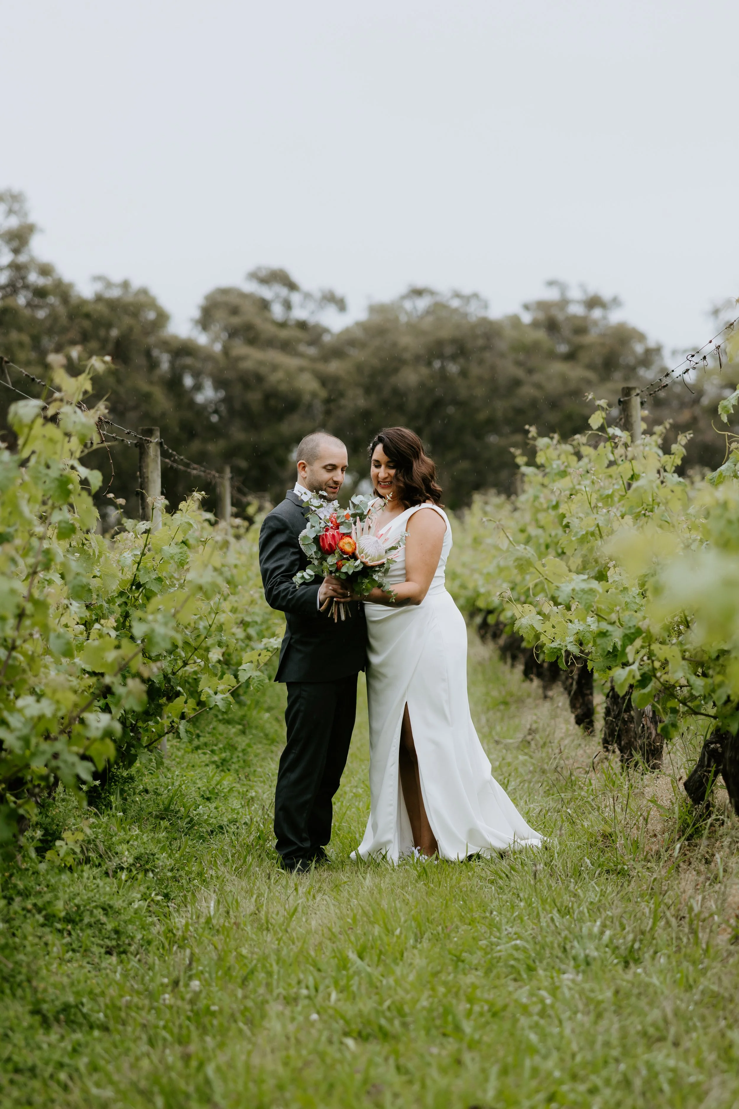 A bride and groom standing in a vineyard, smiling and looking at each other, with the bride holding a bouquet of flowers. The bride is wearing a white wedding dress with a slit, and the groom is in a black suit. There are green grapevines and trees in the background.