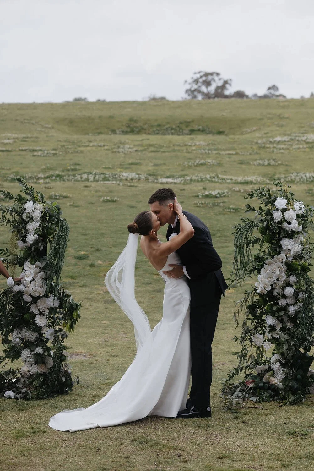 A bride and groom kiss at an outdoor wedding ceremony during the day, surrounded by white floral arrangements.