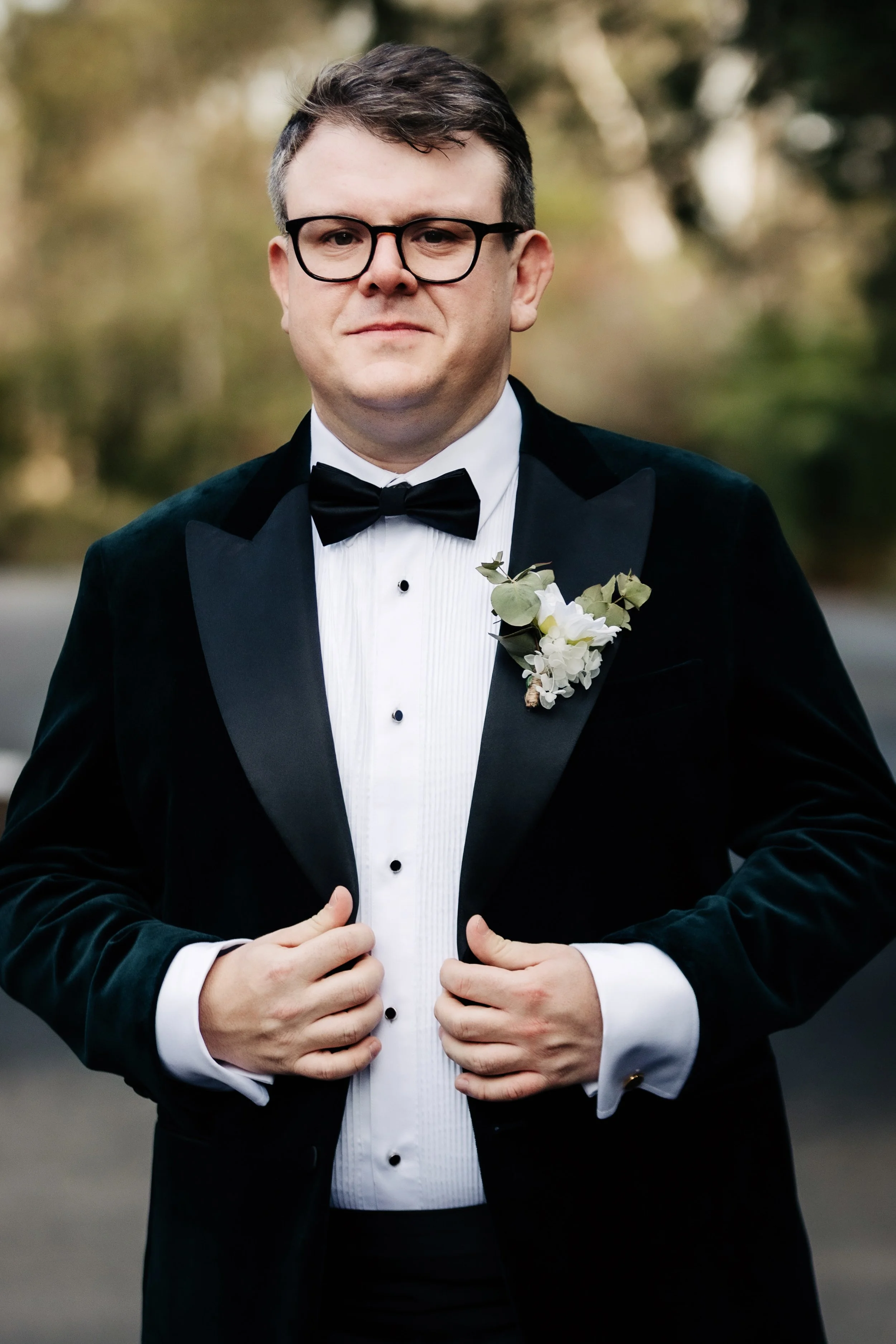 A groom in a tuxedo with a boutonnière, adjusting his jacket, outdoors with blurred trees in the background.