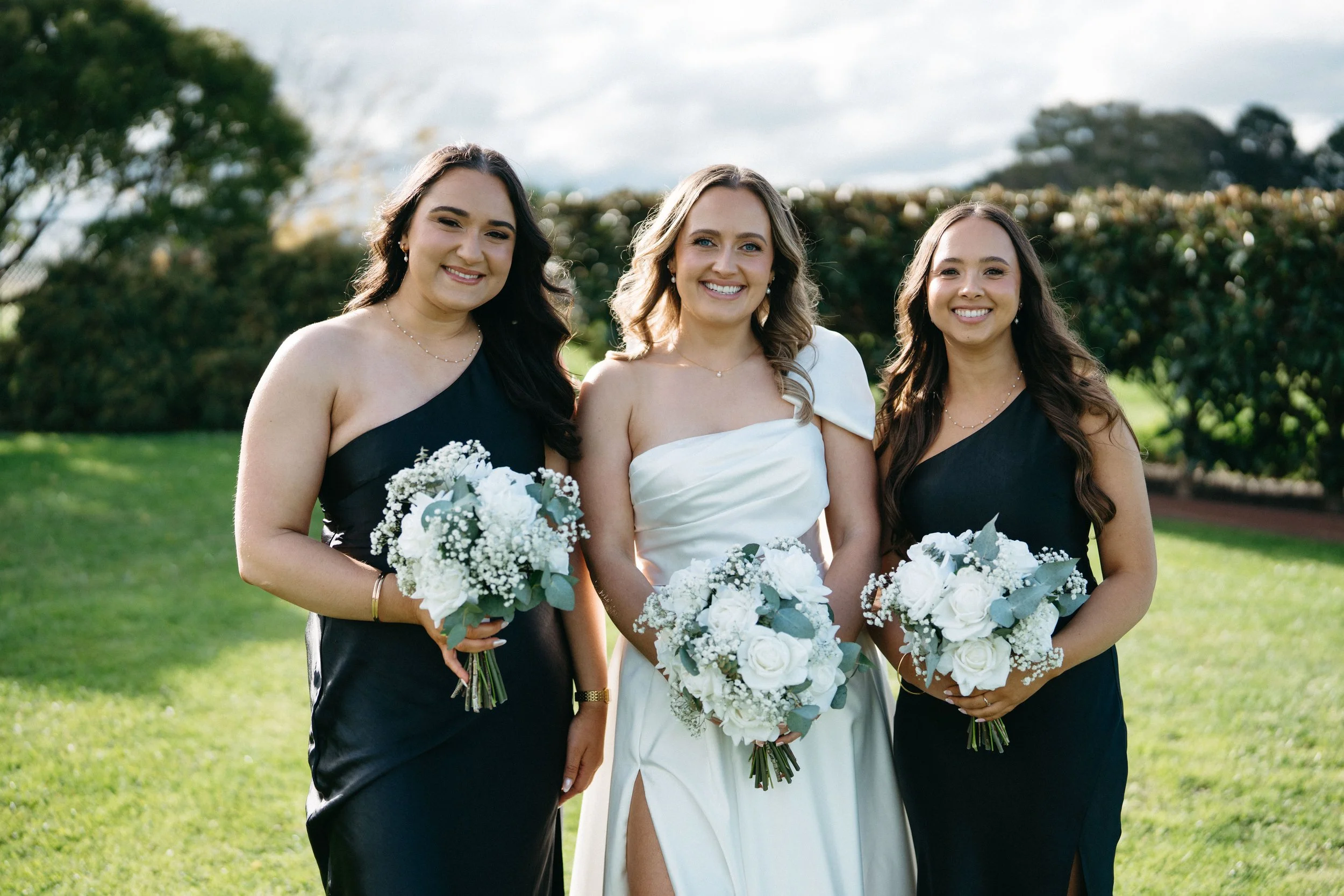 Three women at a wedding, two bridesmaids in black dresses and a bride in a white dress, holding bouquets of white flowers, standing on a grassy outdoor area with trees in the background.