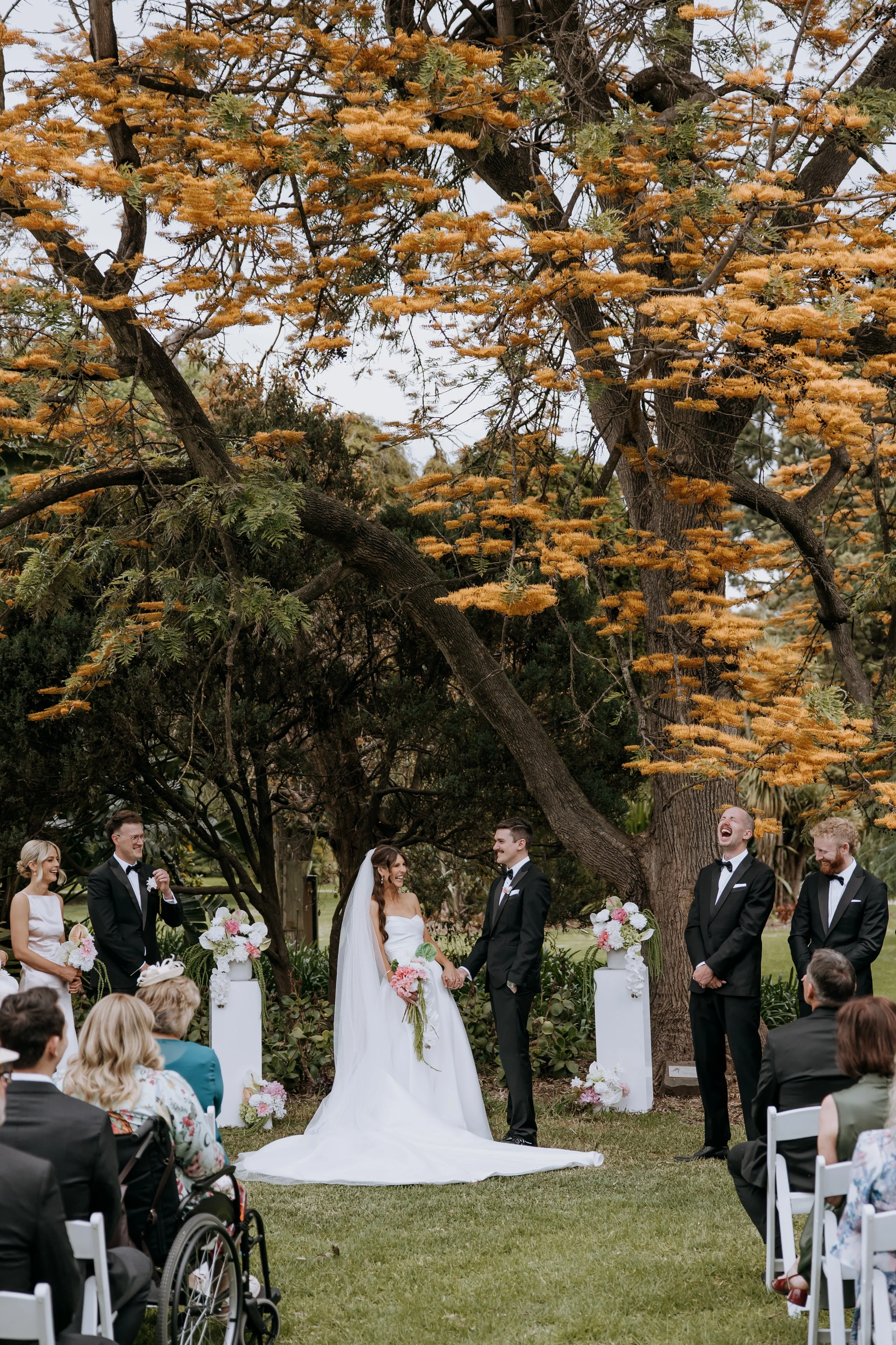 A wedding ceremony outdoors under a large tree with orange leaves, featuring a bride and groom holding hands and smiling at each other, surrounded by wedding party and guests.