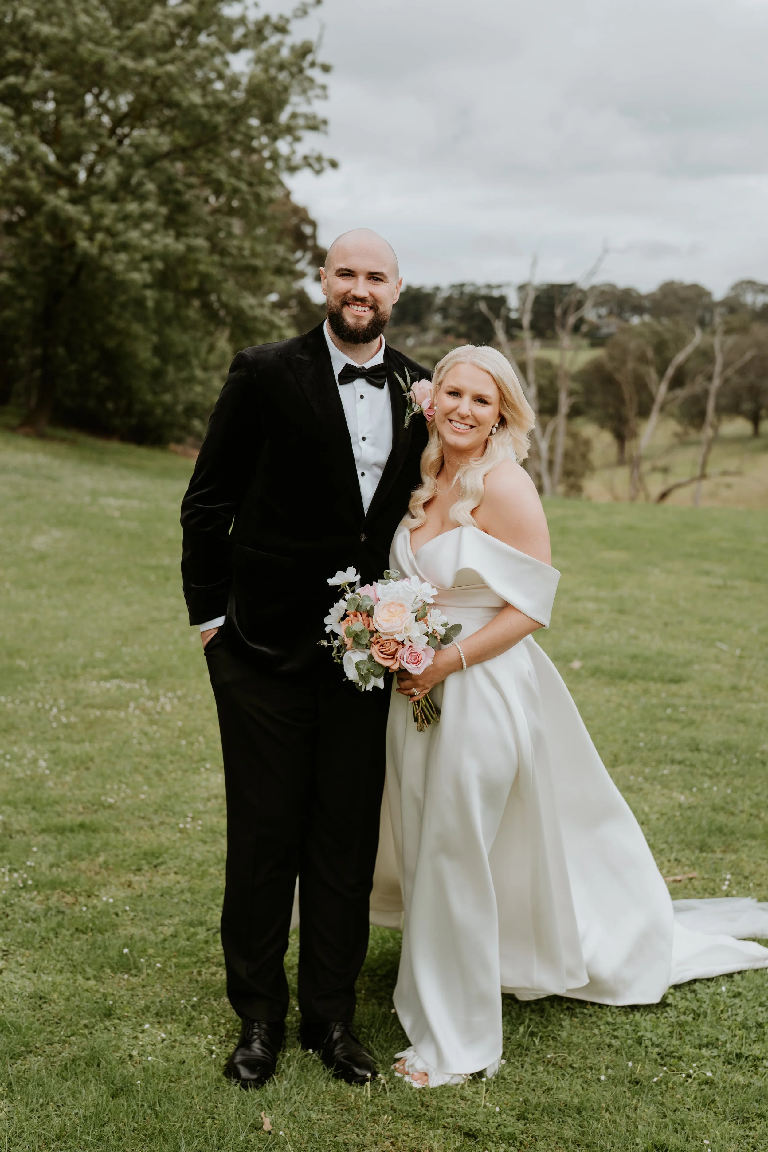 A bride and groom standing together outdoors, the bride holding a bouquet of roses and wearing an off-shoulder white wedding gown, and the groom dressed in a black tuxedo with a bow tie, on a grassy area with trees and cloudy sky in the background.