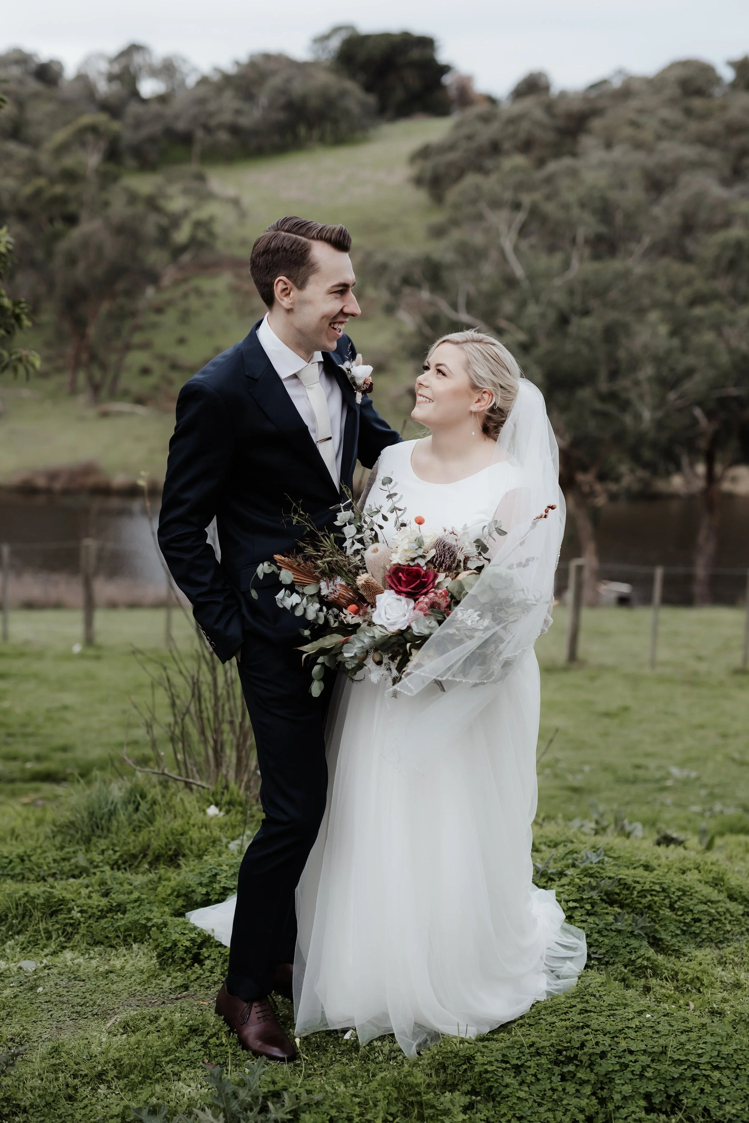 A bride and groom standing outdoors by a fence, smiling at each other, with a lake and trees in the background. The bride is holding a bouquet of flowers and wearing a white wedding dress and veil, while the groom is dressed in a dark suit.