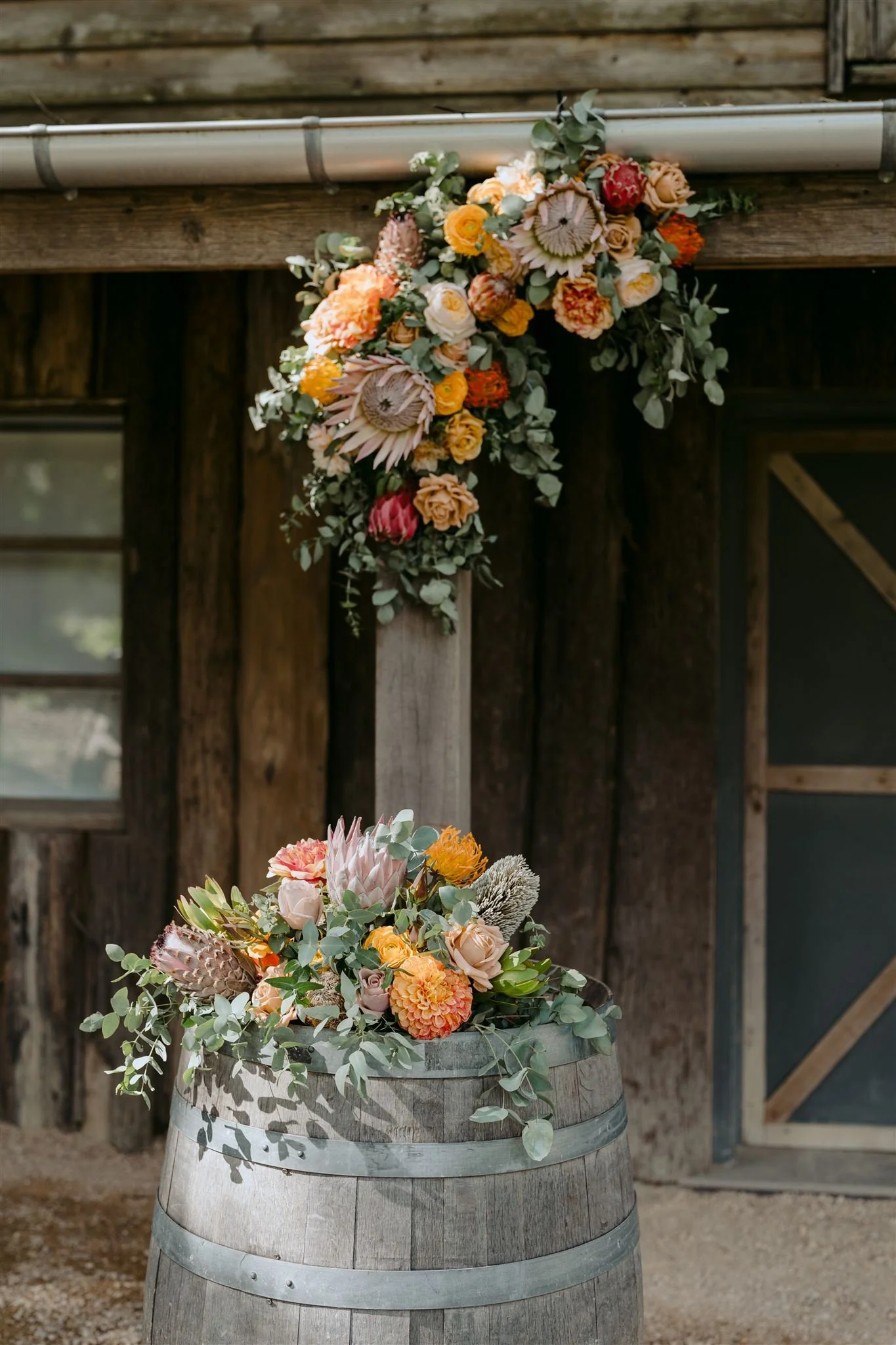 Floral arrangement with various flowers including protea, roses, and protea on a wooden barrel, with a rustic barn wall in the background.
