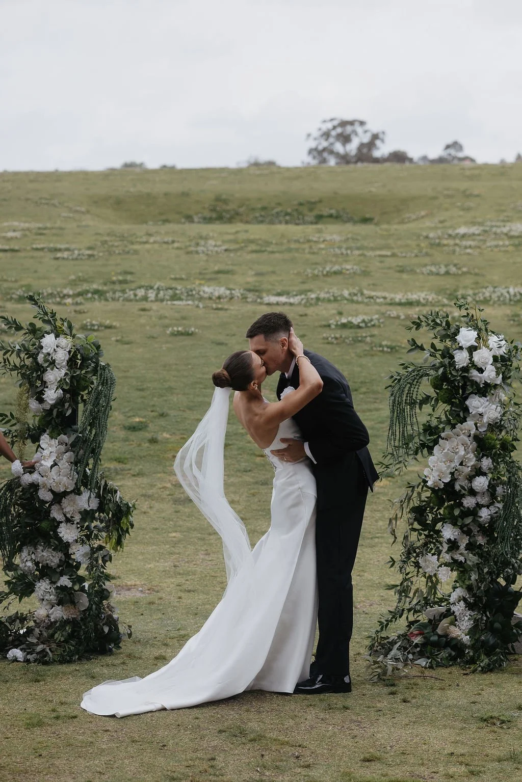 A bride and groom sharing a kiss during their wedding ceremony outdoors, surrounded by floral arrangements, on a grassy field with trees in the background.