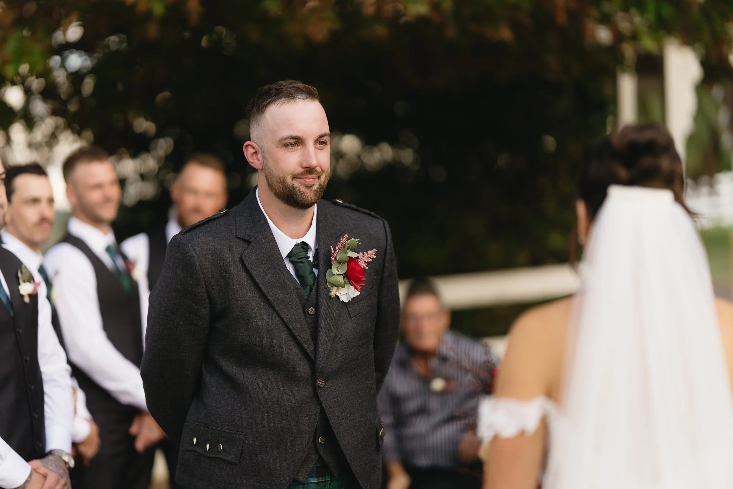 Groom with a beard wearing a dark suit, green tie, and boutonniere, during his wedding ceremony outdoors, facing the bride with her veil, with groomsmen standing in the background.
